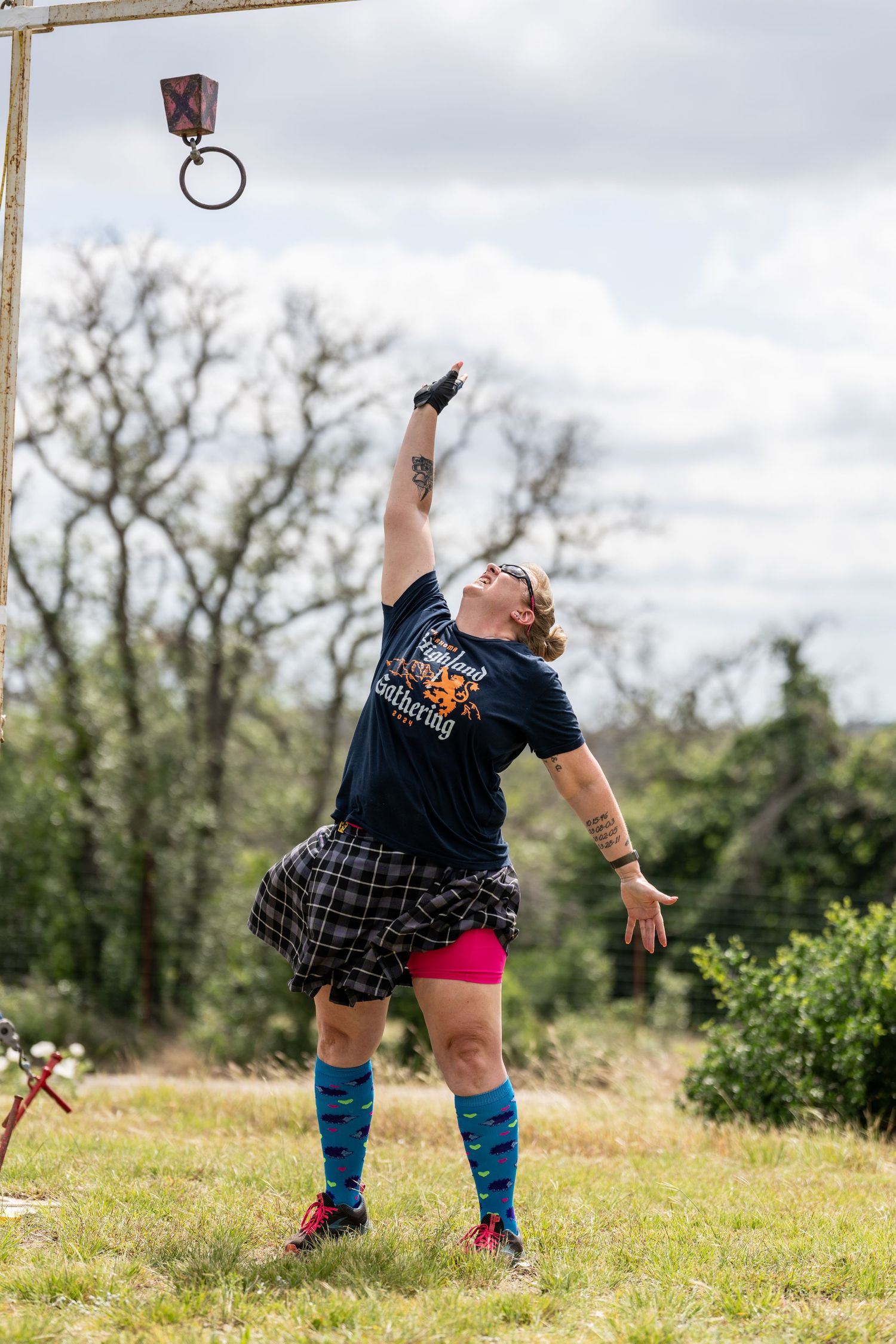 Woman in kilt and blue socks reaches up during Highland Games competition outdoors.