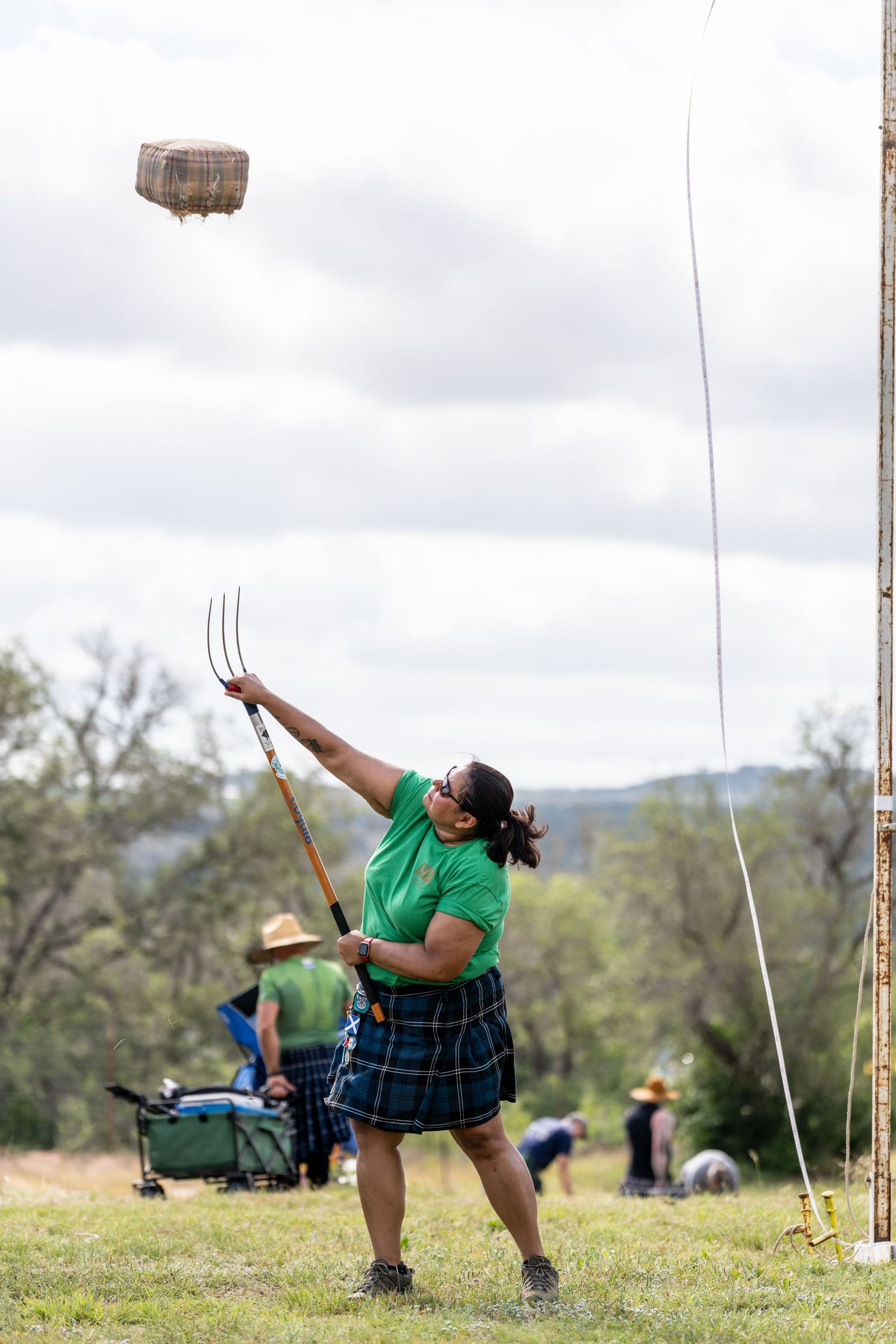 Woman in green shirt and tartan kilt throws sheaf with pitchfork at Highland Games competition outdoors.