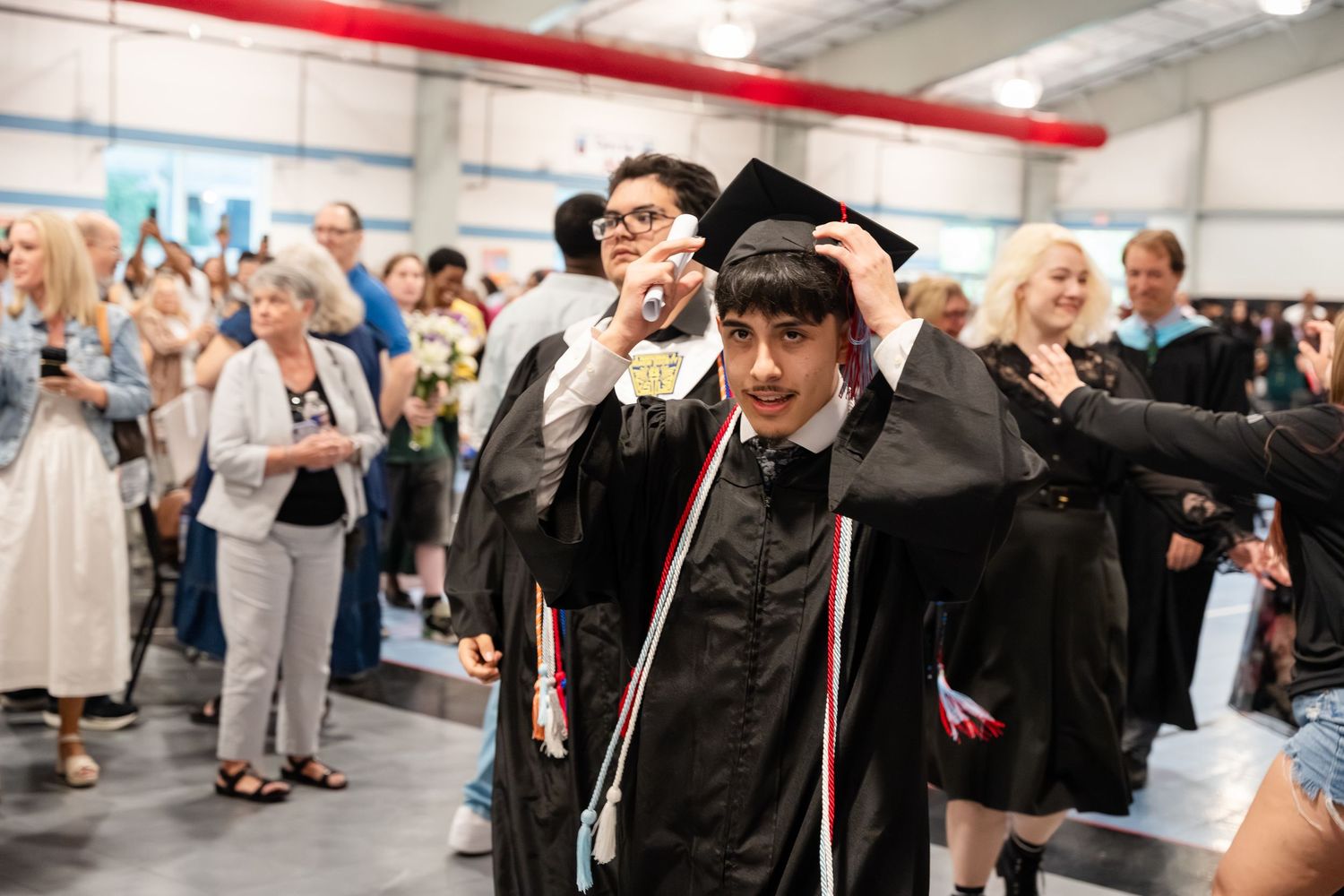 Graduates in black caps and gowns celebrate at an indoor graduation ceremony surrounded by cheering family members.