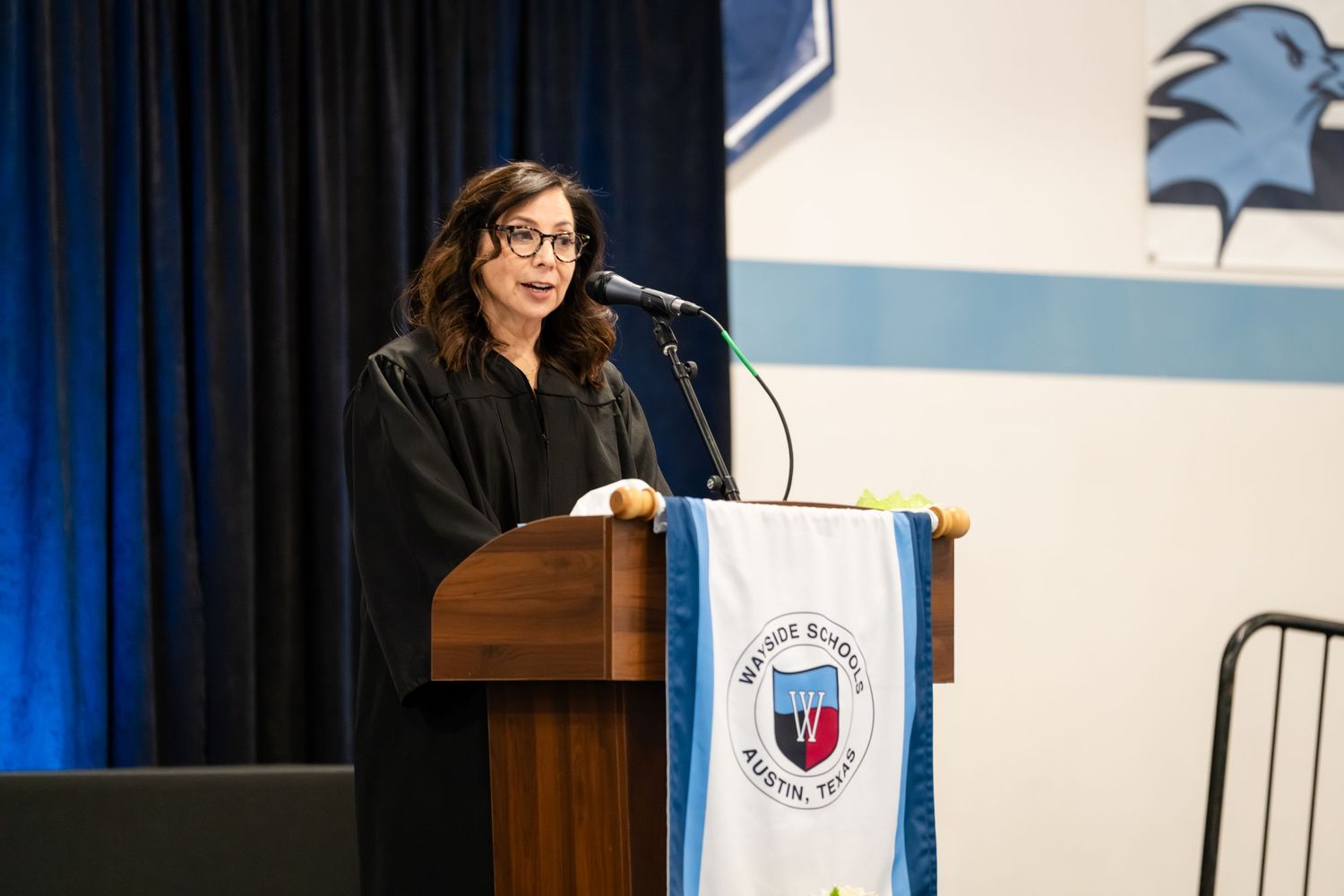 Woman in academic regalia speaking at a podium during a graduation ceremony in a school gymnasium.