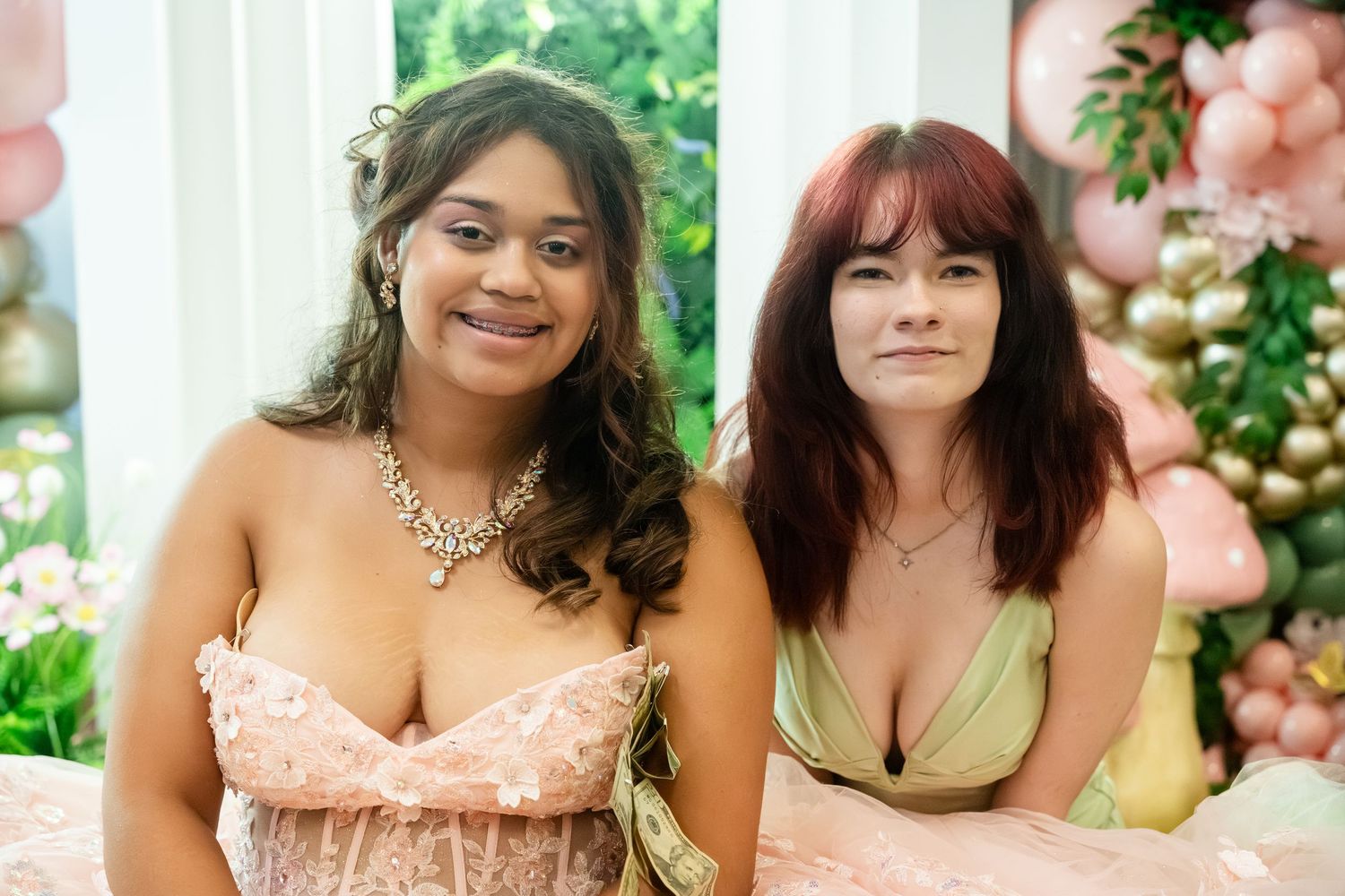 Two young women in elegant formal dresses posing together at a decorated event with balloon backdrop.