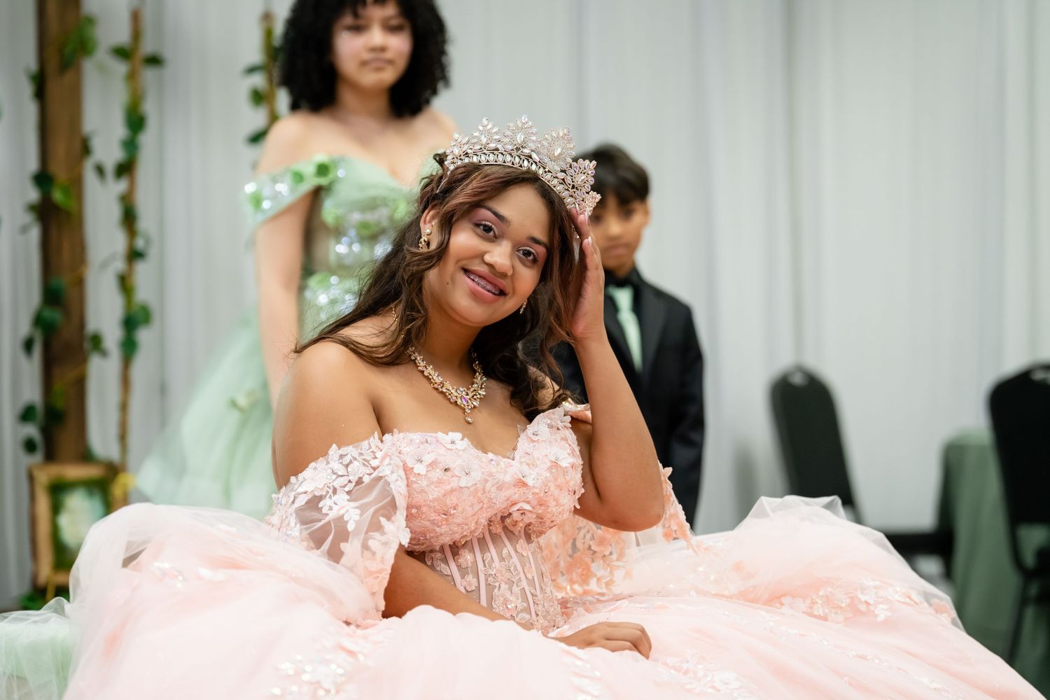 Young woman in pink ball gown and tiara poses gracefully at a quinceañera celebration.