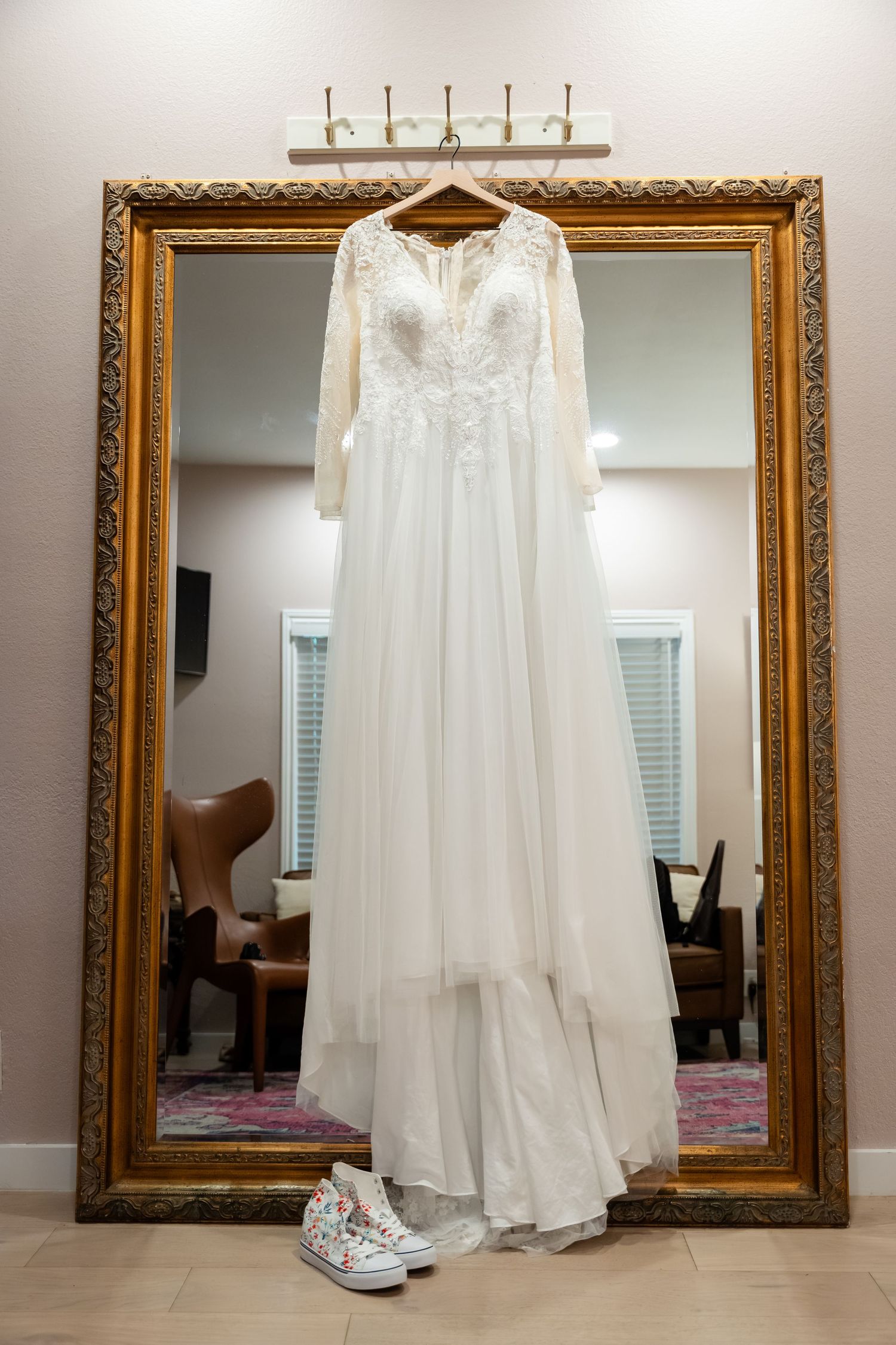 White lace long-sleeve wedding gown hanging on ornate gold mirror with floral Converse sneakers below.