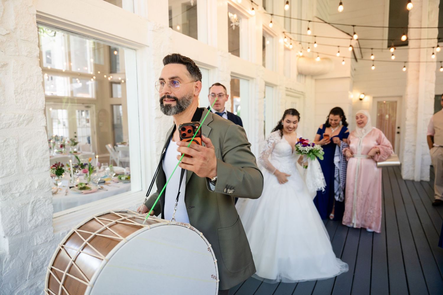 A musician plays a drum leading a wedding procession with a bride and guests in a decorated venue.