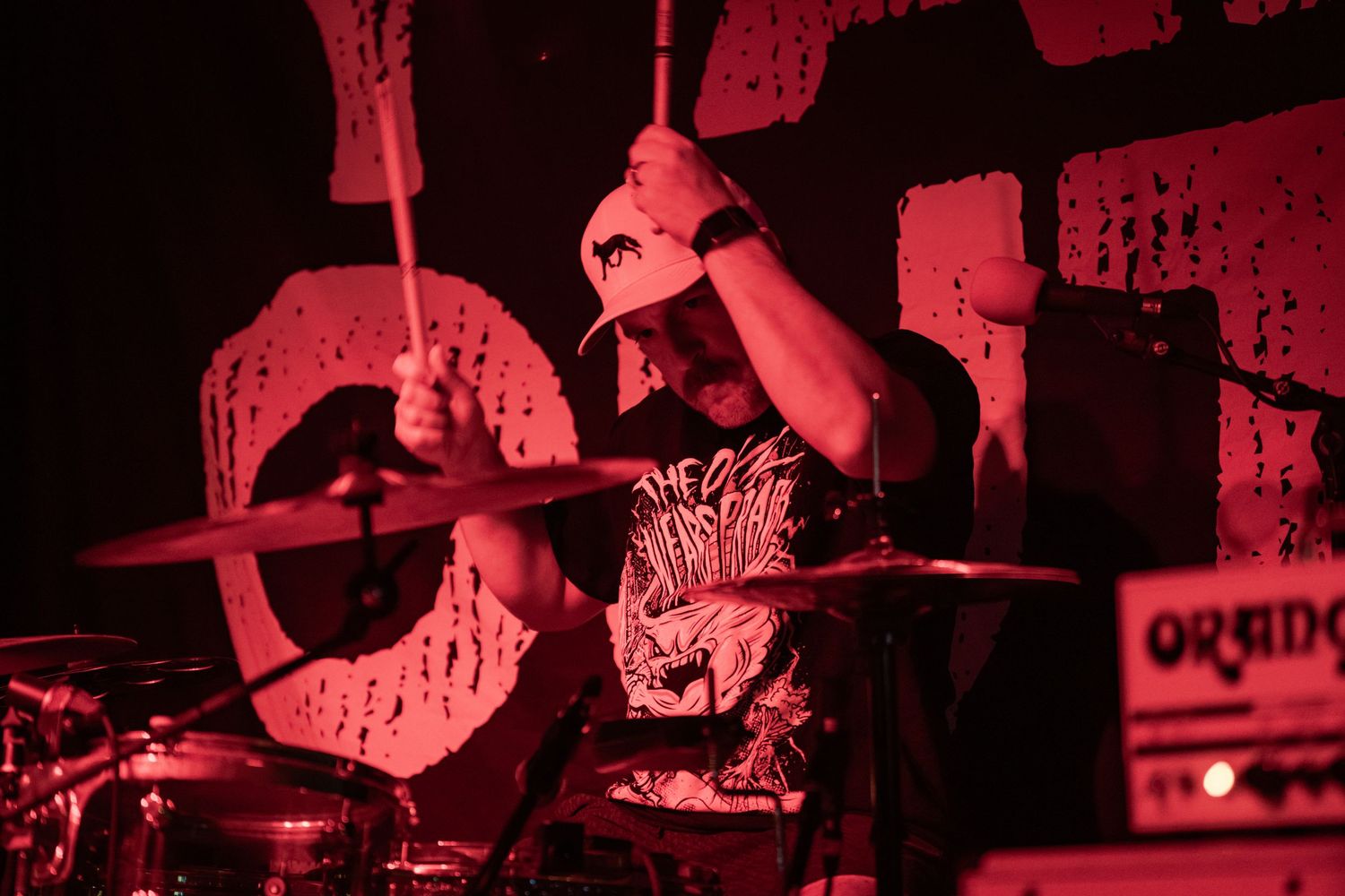 Drummer performing intensely on stage under red lighting at a live music concert.