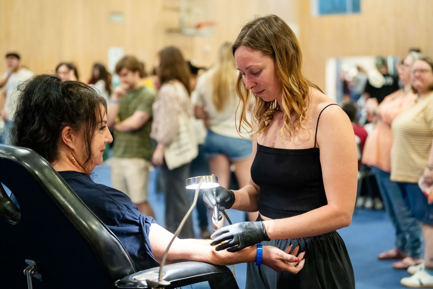 Healthcare worker in black dress draws blood from seated patient at busy medical donation event.