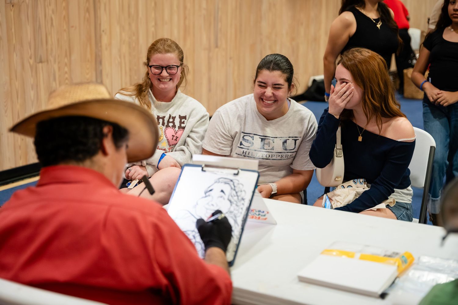 Three women laugh joyfully while a man in a cowboy hat signs autographs at an indoor event table.