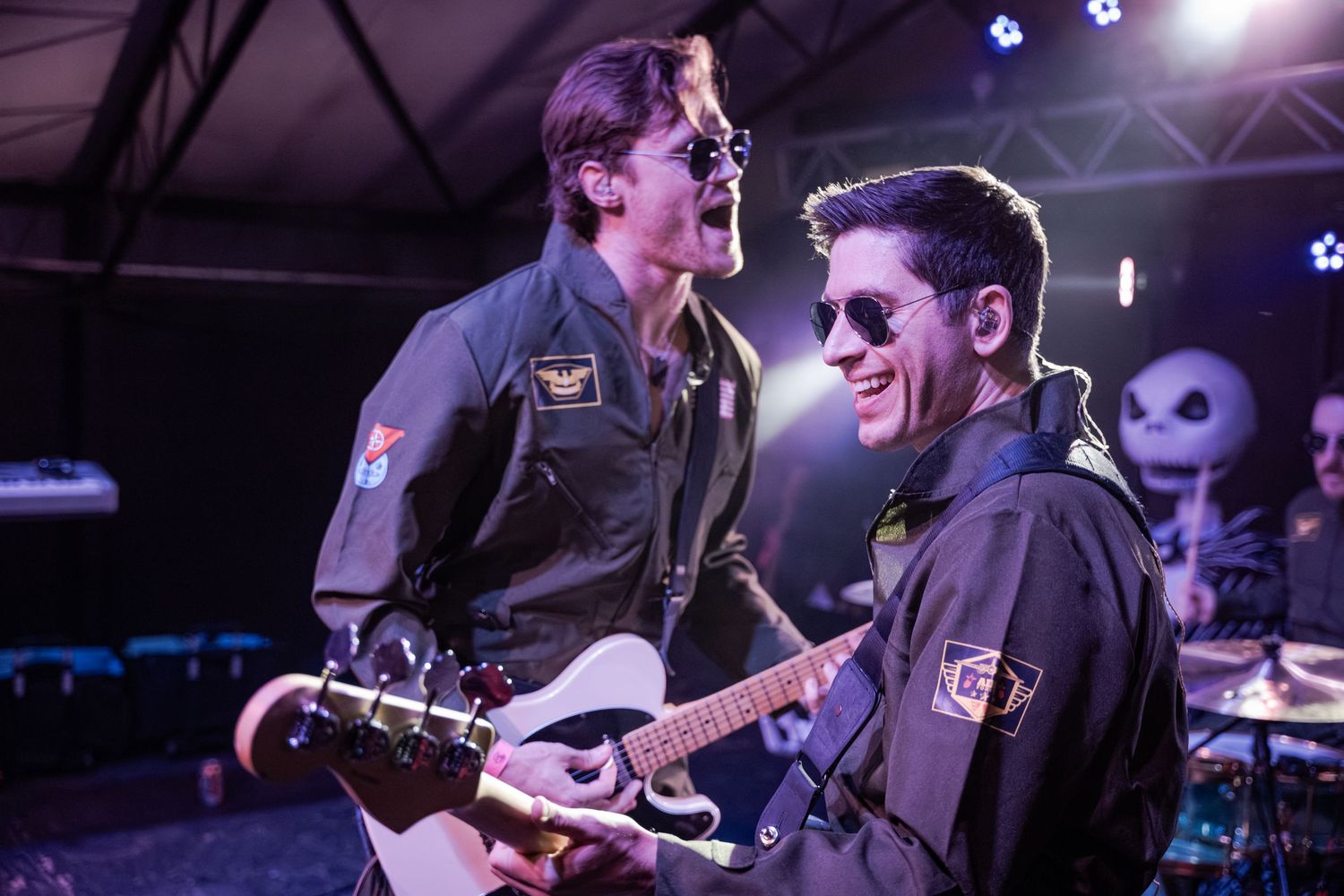 Musicians perform on stage with guitars and microphones under purple stage lighting during a live concert performance.