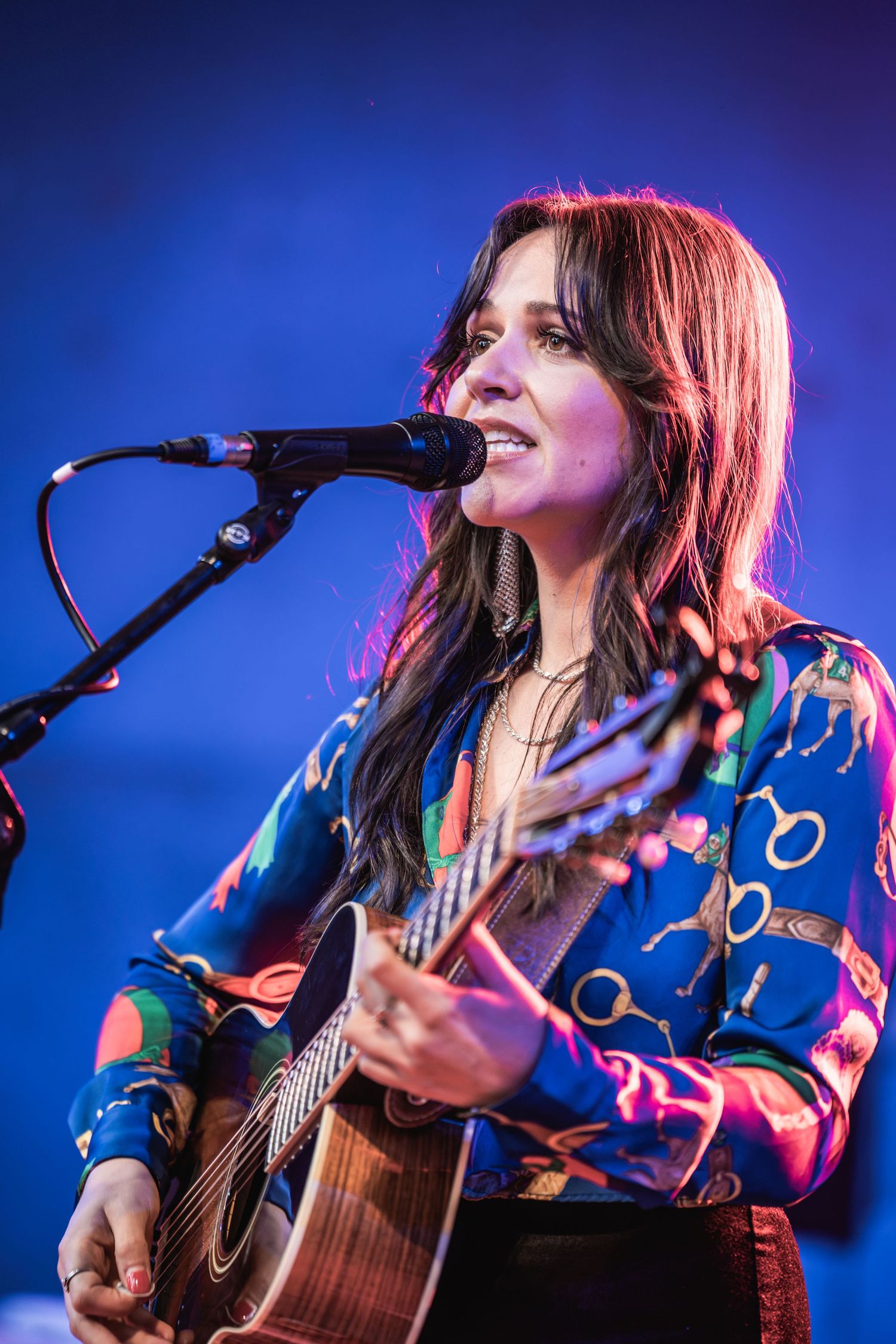 A musician performs on stage with an acoustic guitar against a blue-lit backdrop during a live concert performance.