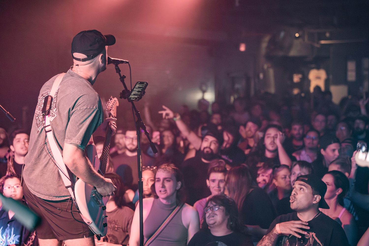 A performer on stage energetically entertains a packed crowd at a concert venue with moody red lighting.