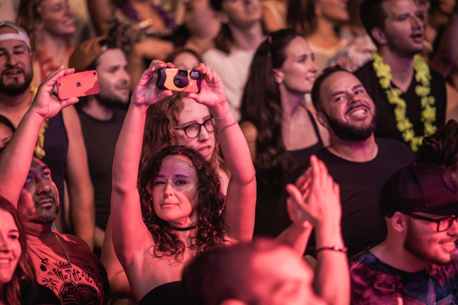 Concert crowd enjoying a show with red lighting as audience members record on phones and smile in excitement.