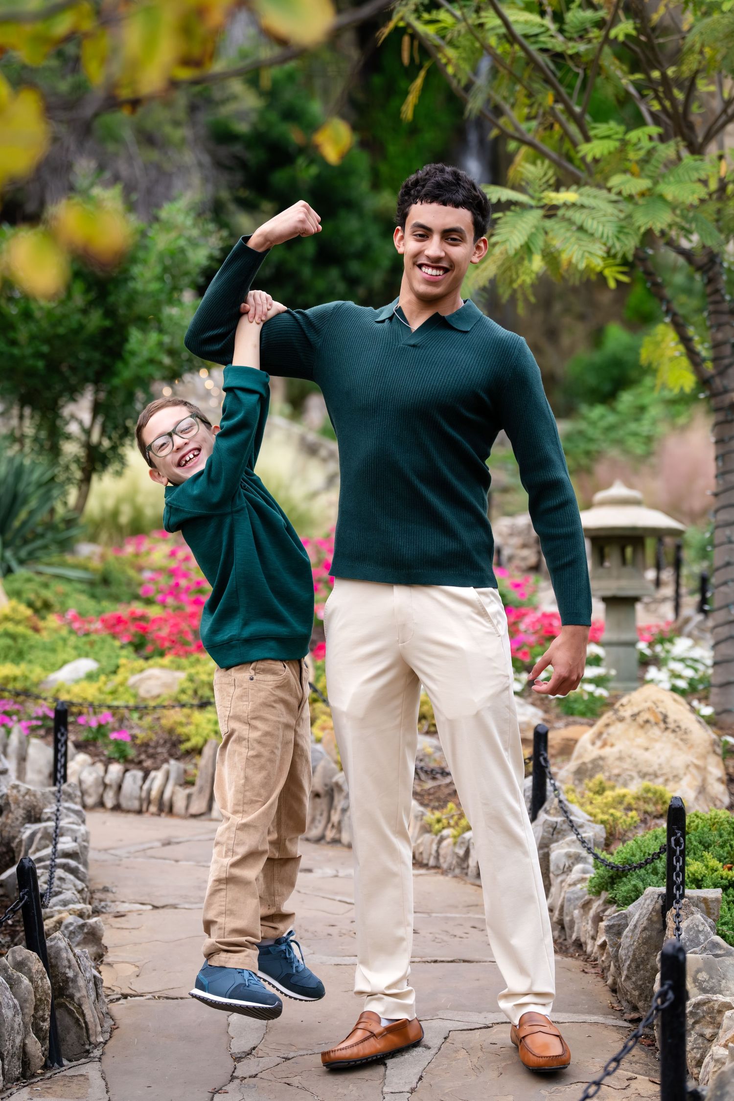 Two people in matching green sweaters pose playfully on a garden path with stone borders and colorful flowers.