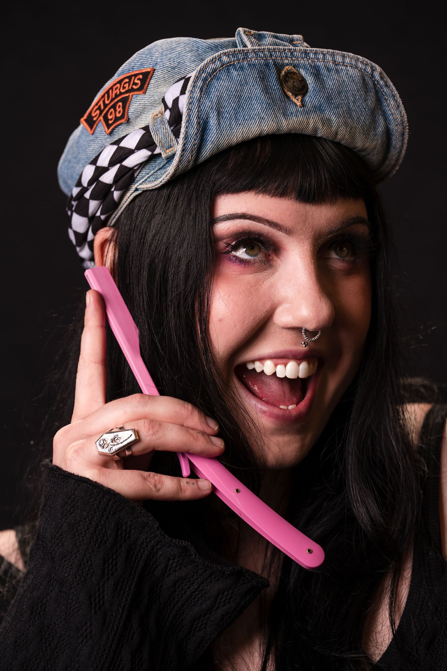 Alternative style portrait with denim cap featuring checkered patches, nose ring, and pink plastic phone against dark background.