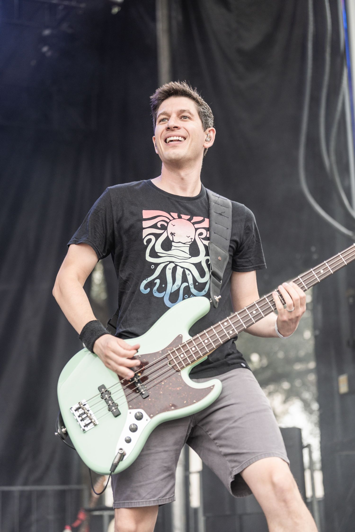 Bass player in black t-shirt with octopus design performs energetically on stage with mint green bass guitar.