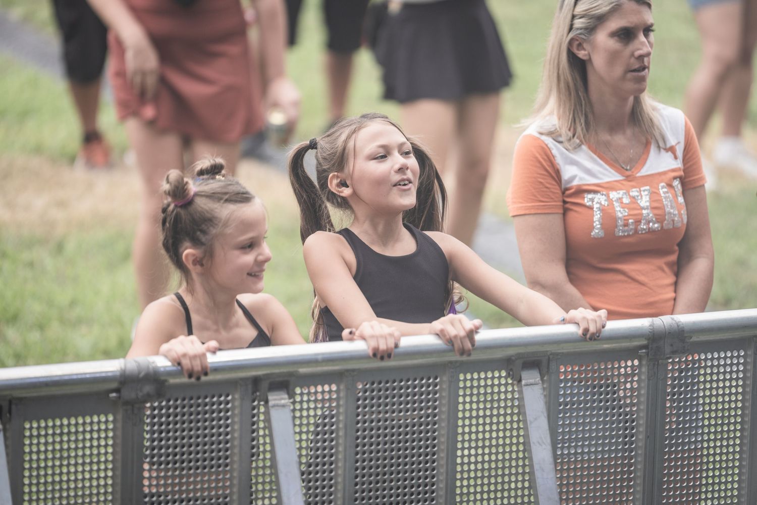 Three spectators lean against a metal railing while watching an outdoor event on a sunny day.