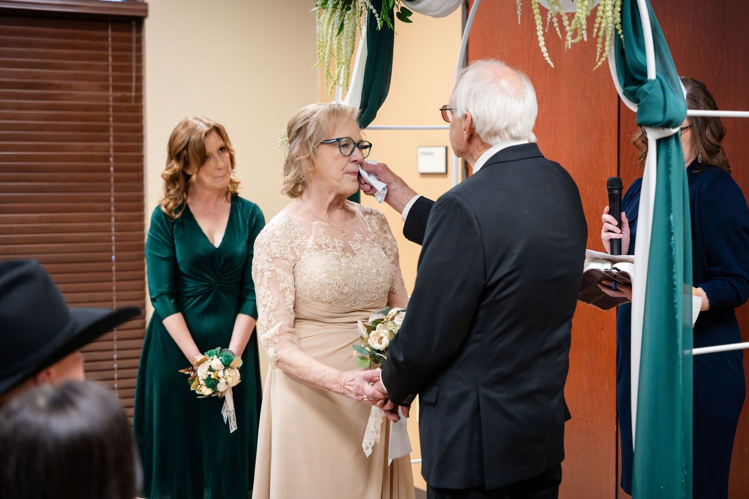 An intimate wedding ceremony takes place under teal drapery with attendants in green and champagne attire.