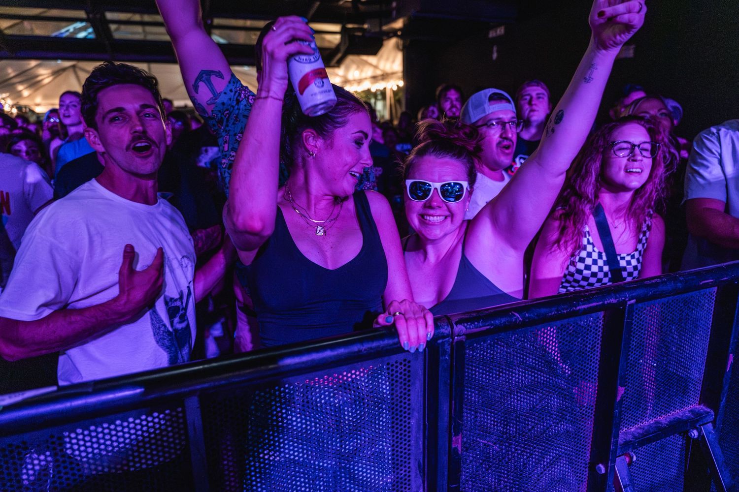 Excited concert crowd cheers behind barrier railing at music show with purple lighting effects.