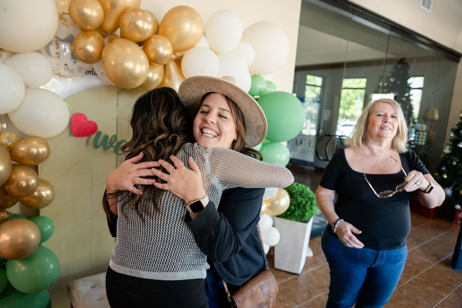 People sharing warm embraces at a celebration with gold and green balloon decorations.