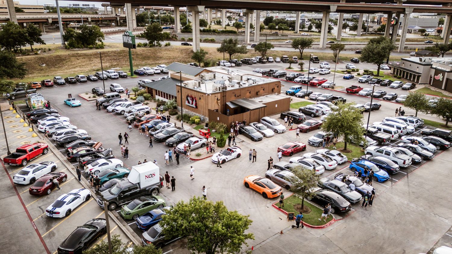 Aerial view of crowded car meet showing parking lot filled with vehicles and attendees.