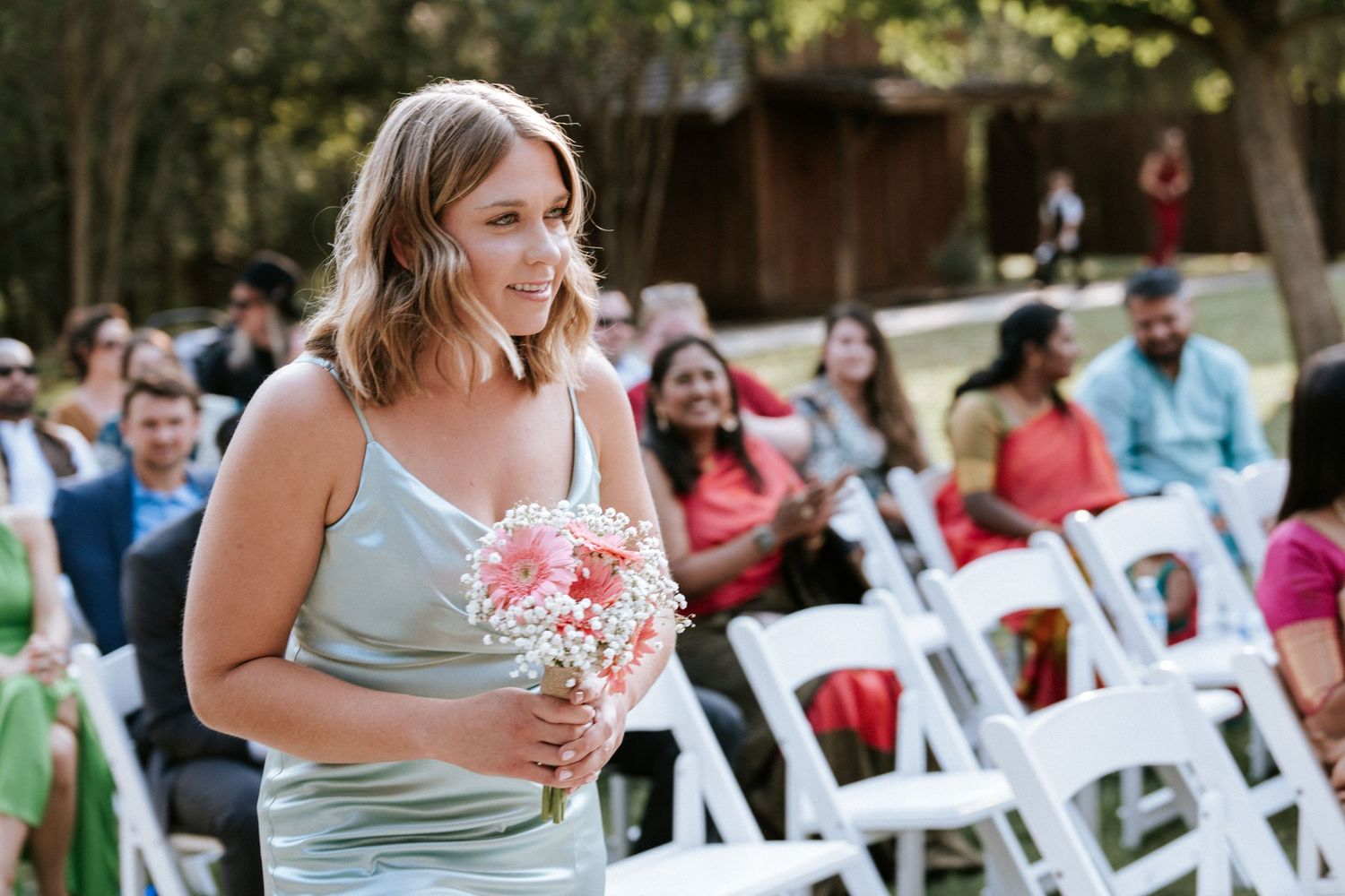 Bridesmaid in mint green dress holding pink bouquet at outdoor ceremony.