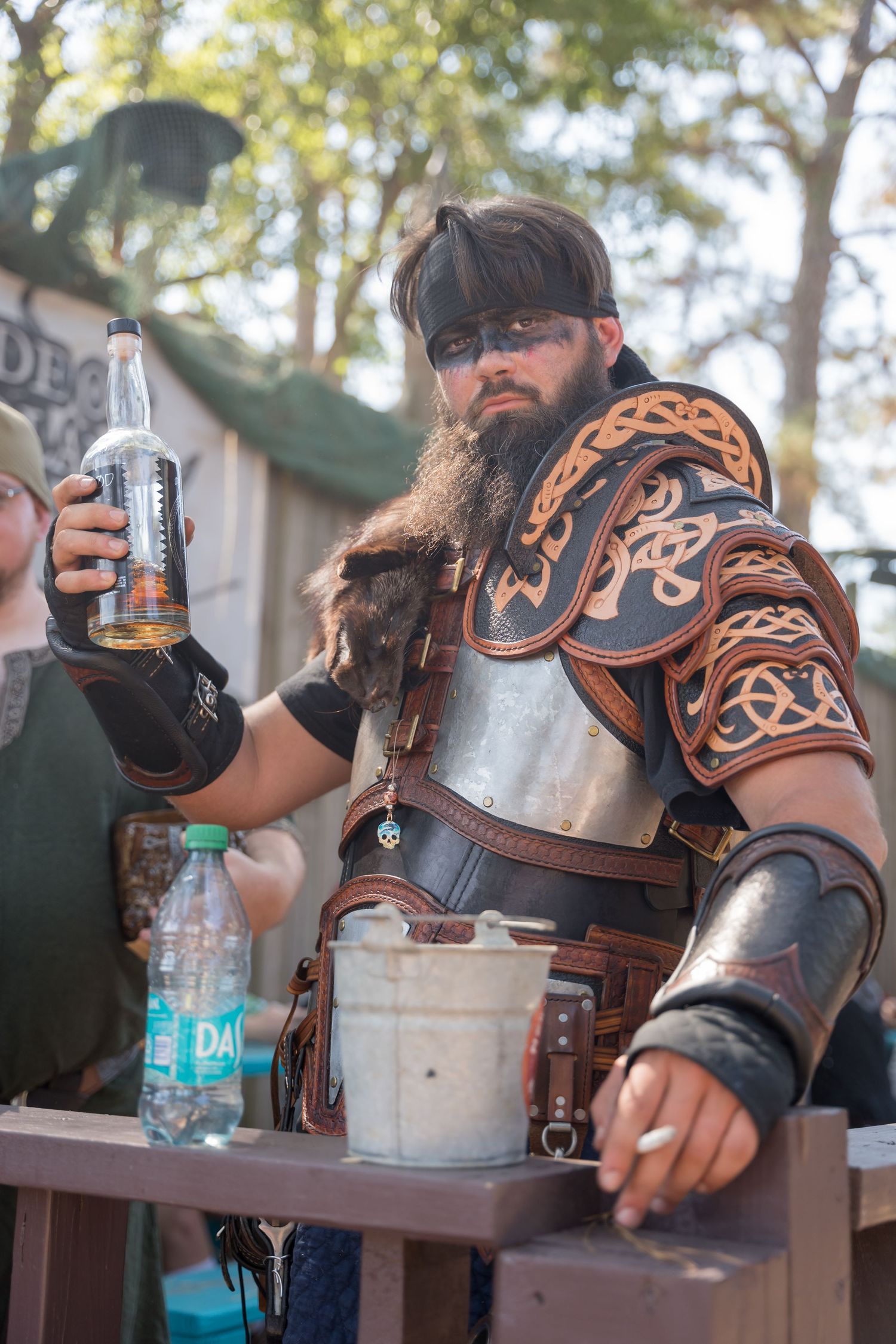 A costumed performer in leather armor and beard holds a bottle while working at an outdoor medieval fair.