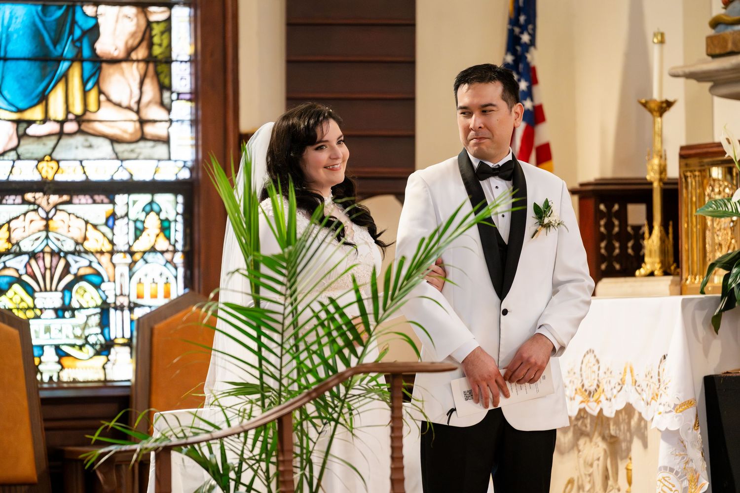 A couple stands together during their wedding ceremony in a church with stained glass windows and palm decor.