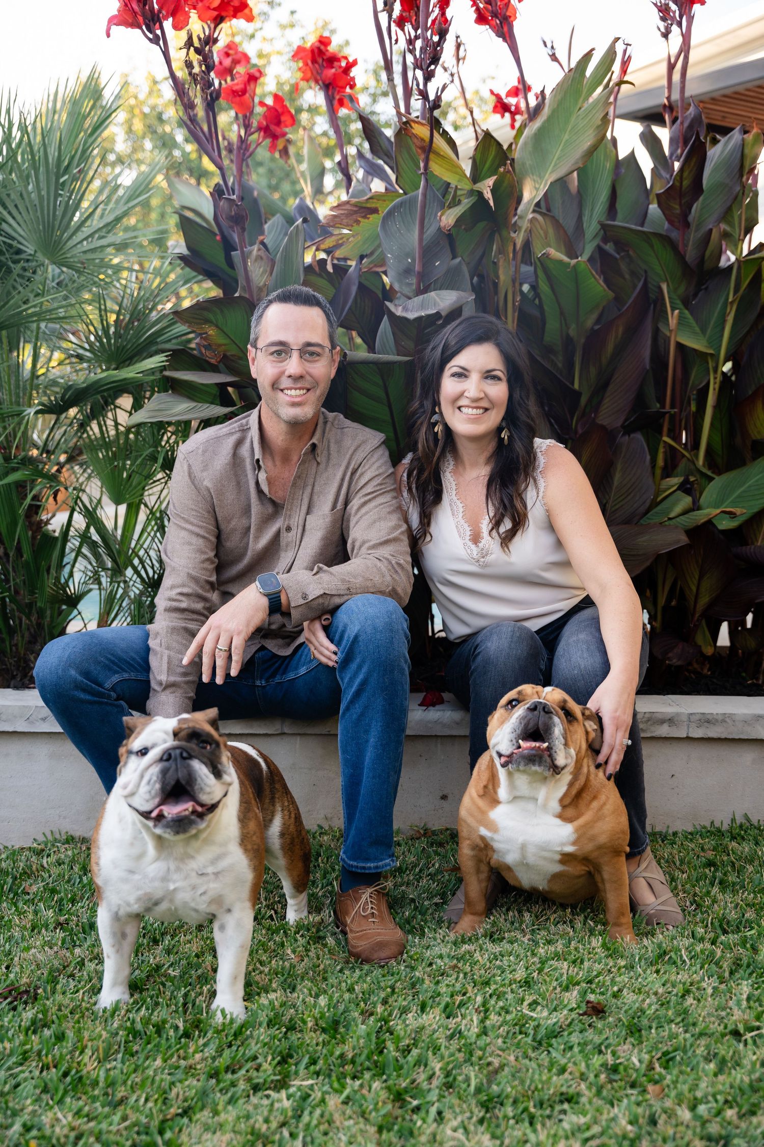A couple sits with their two English Bulldogs in front of red flowering plants in a garden setting.