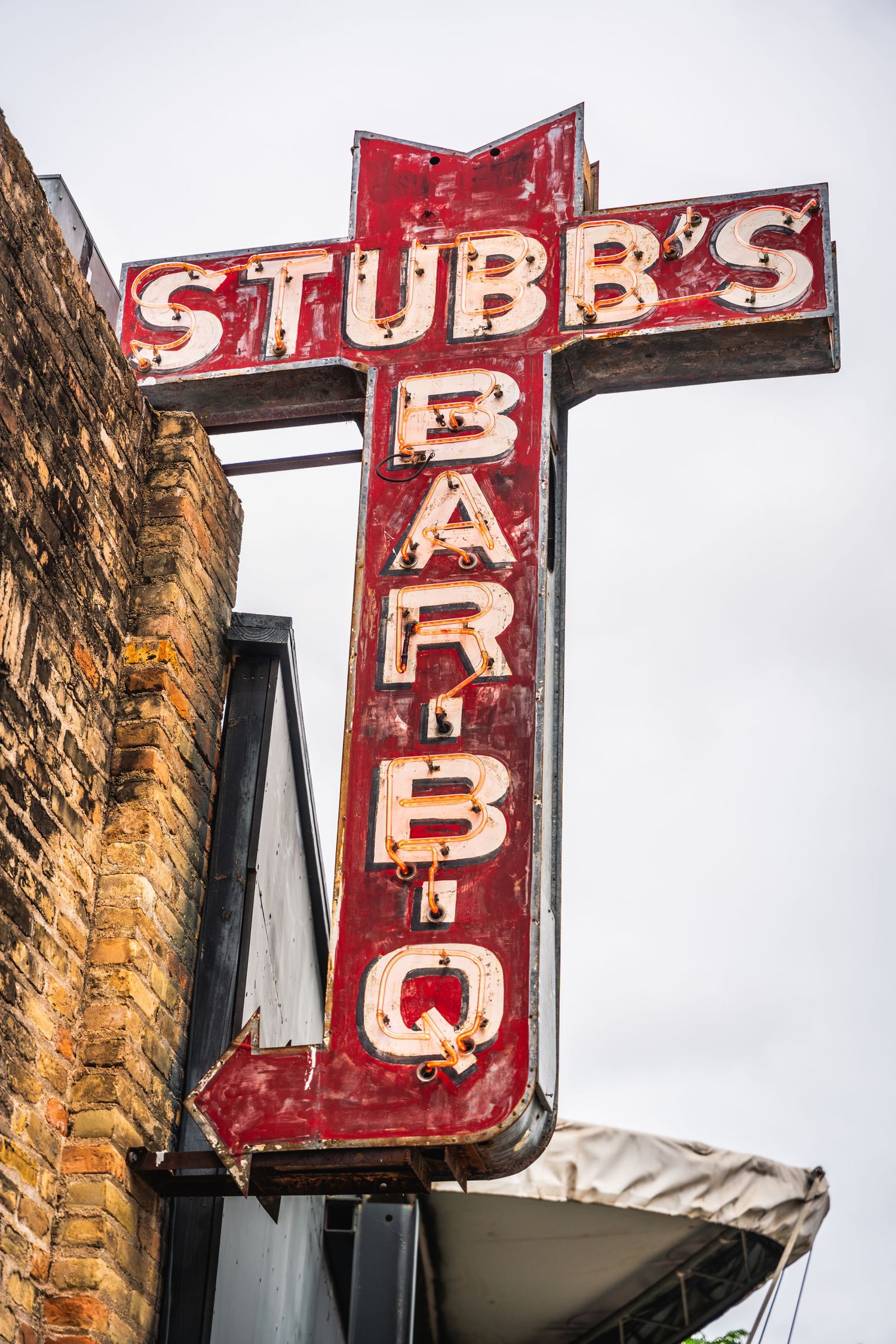 Iconic red neon Stubb's BBQ sign mounted on brick building in cross shape against cloudy sky.