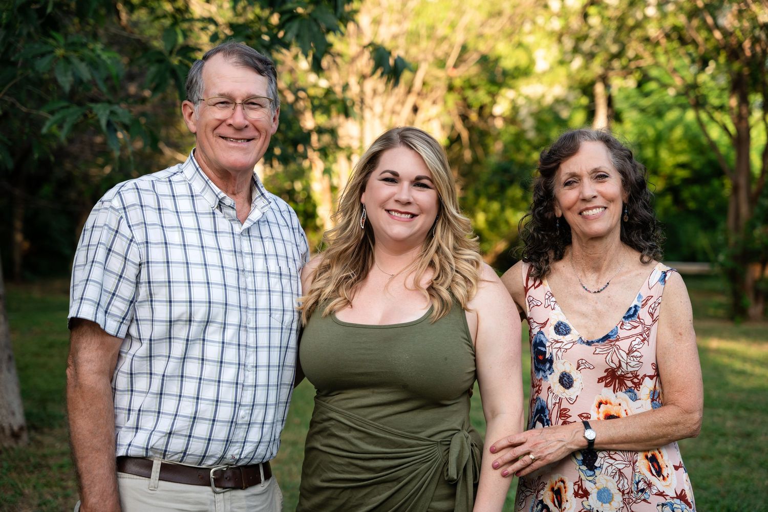 Three people pose casually outdoors for family photo with green foliage backdrop.
