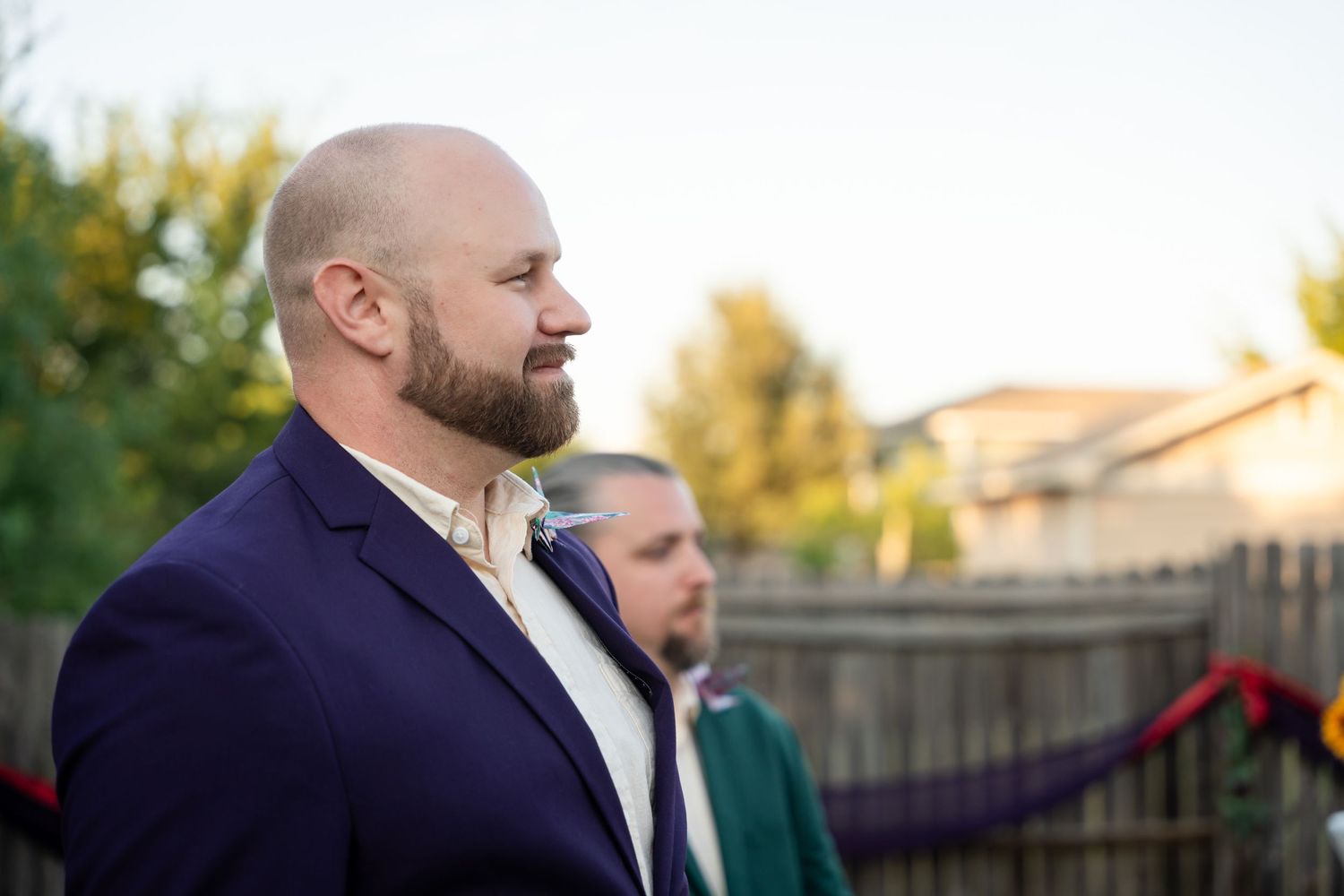 A person in a navy blazer stands outdoors during a wedding ceremony at sunset.