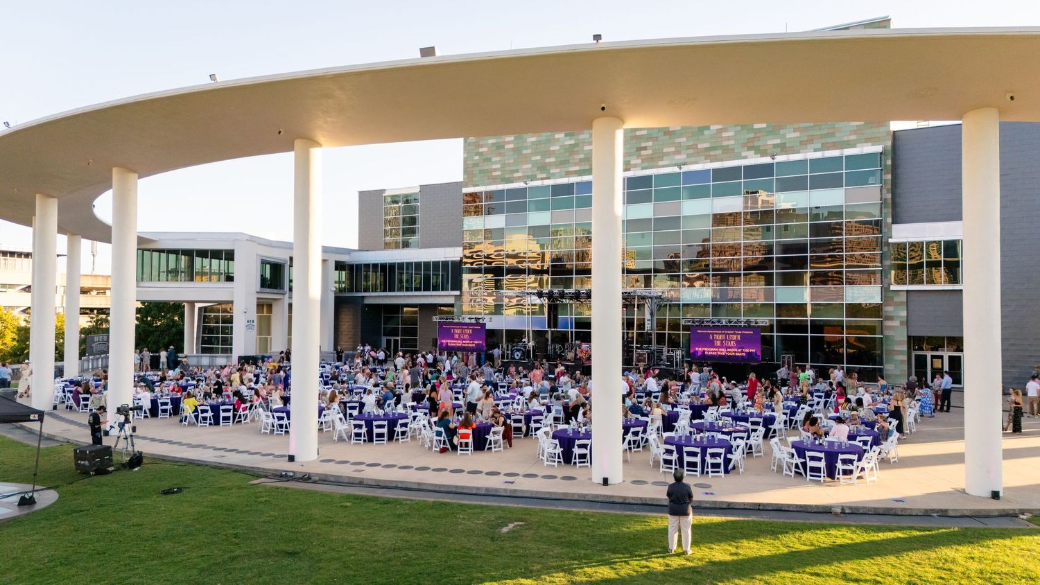 Large crowd gathered under covered outdoor venue area with modern buildings in background.