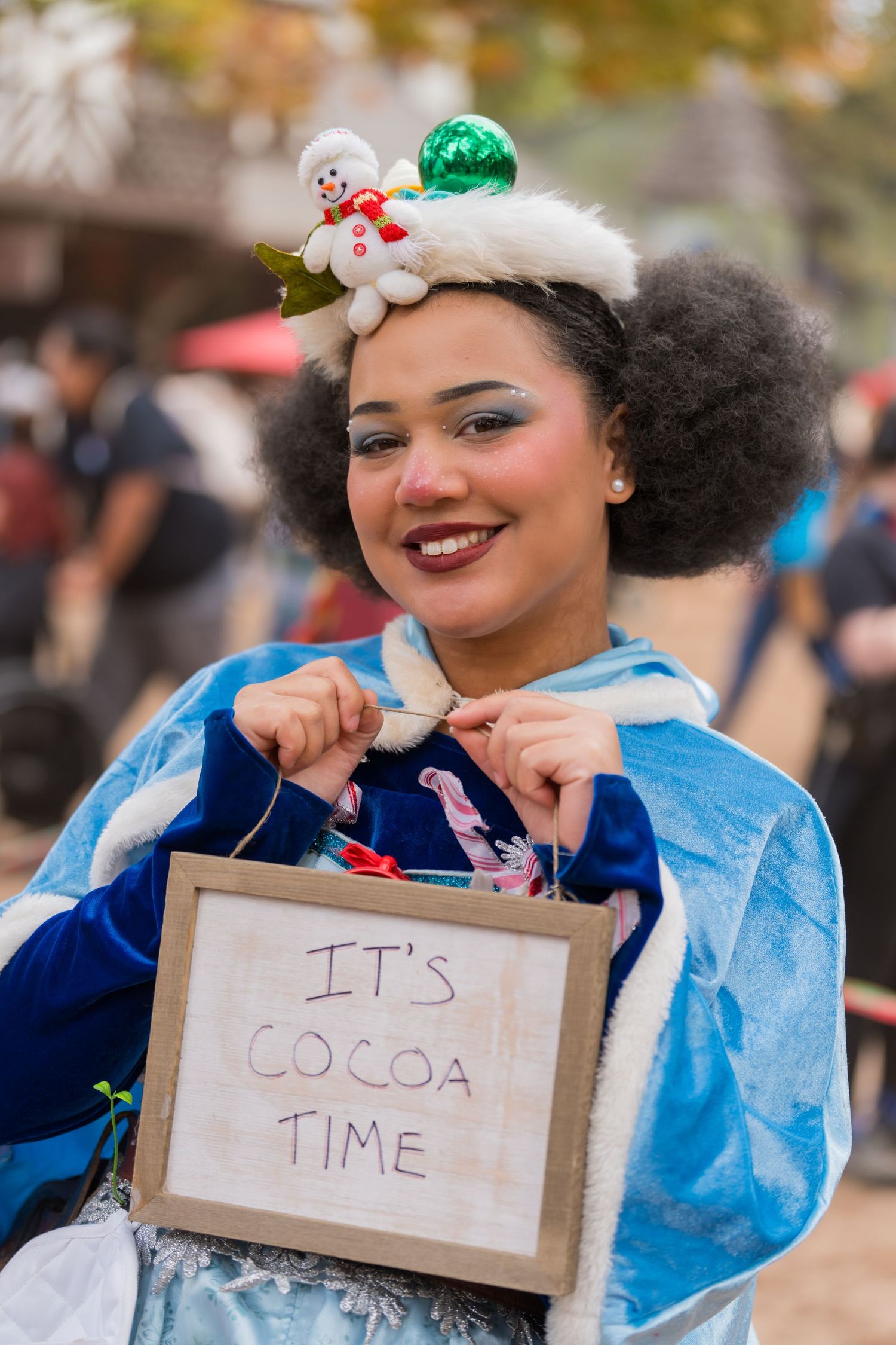 Festival goer wearing blue dress and snowman hair accessory holds sign reading 'It's cocoa time'.