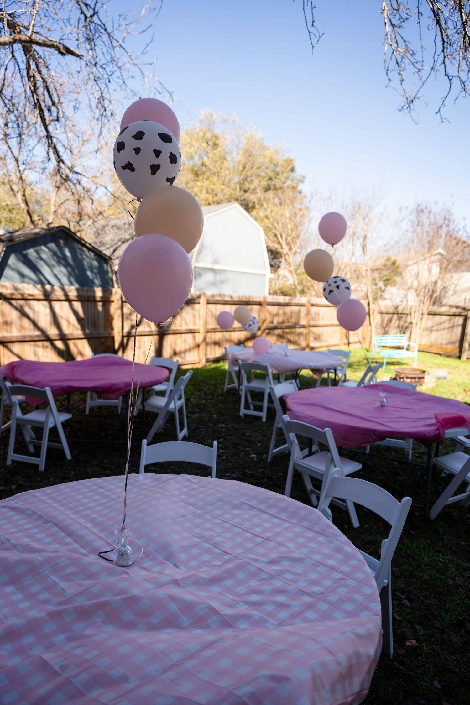 Pink and white outdoor party setup with gingham tablecloths, white folding chairs and floating heart-patterned balloons in a backyard.