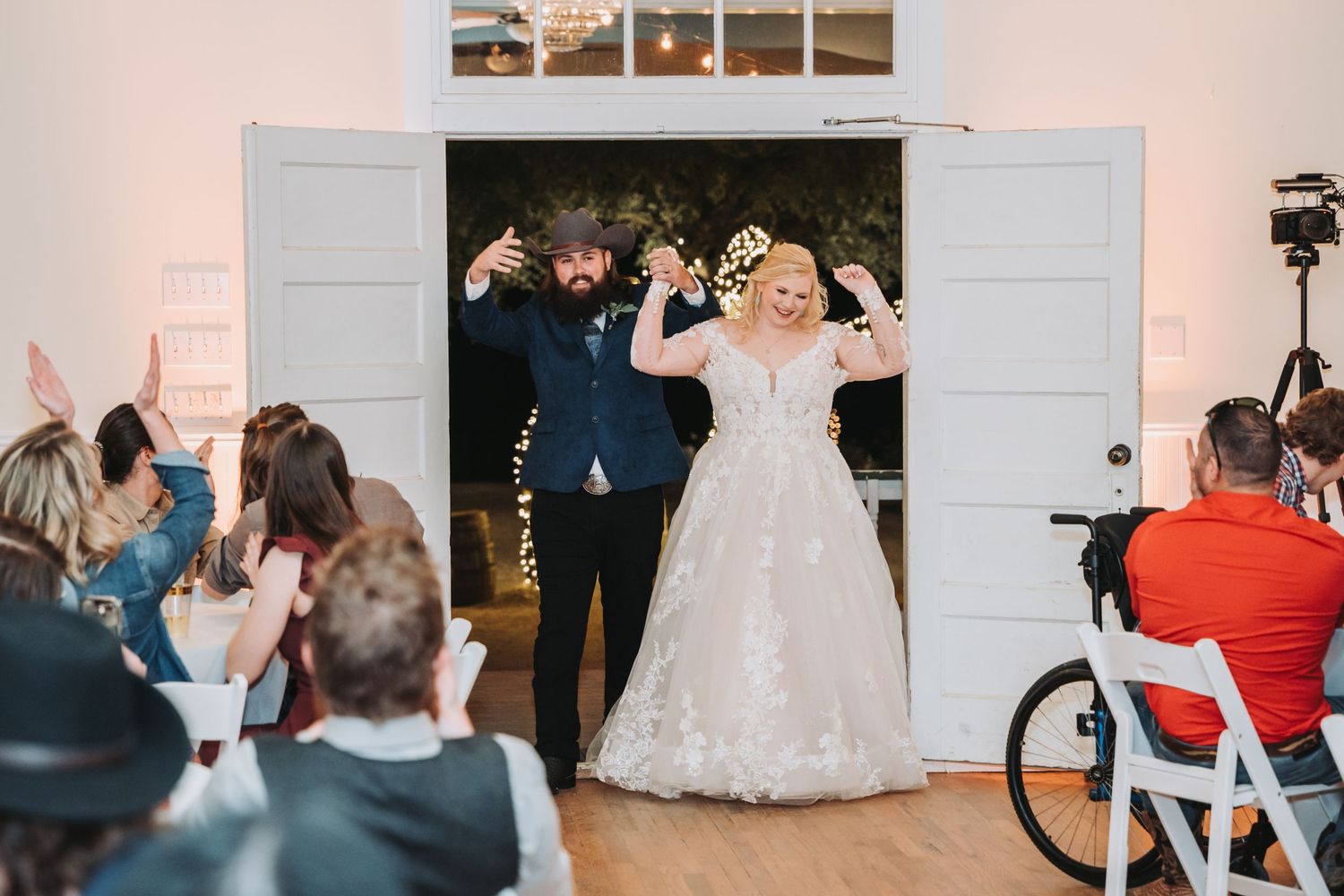 A newlywed couple makes their grand entrance as guests cheer in a white indoor venue.