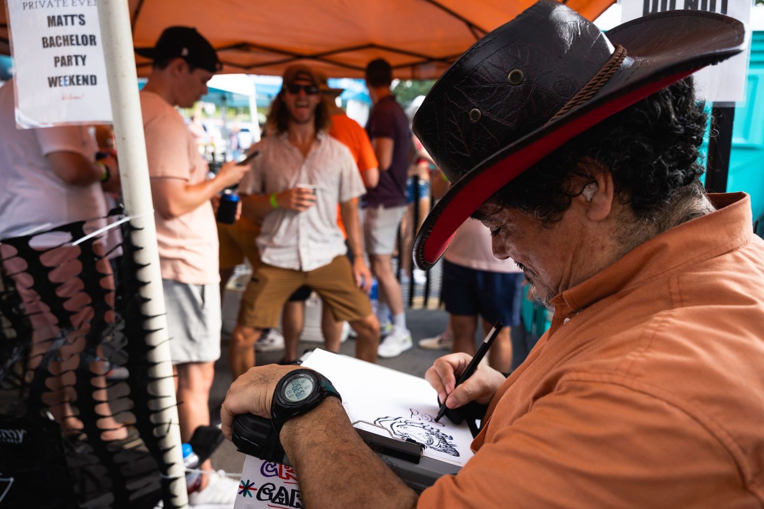 A person in an orange shirt and black hat signs autographs at an outdoor event.