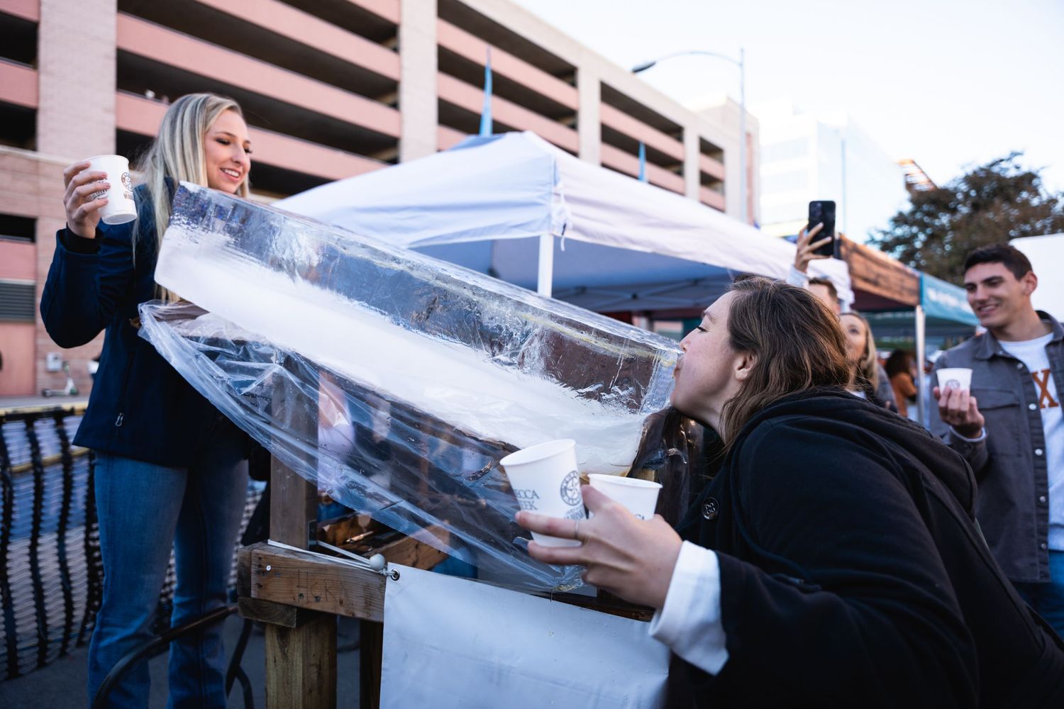 Two people pose while drinking from an ice luge at an outdoor party.
