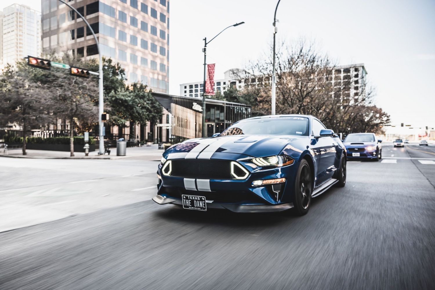 Blue Ford Mustang GT with racing stripes driving on a city street with modern buildings in the background.