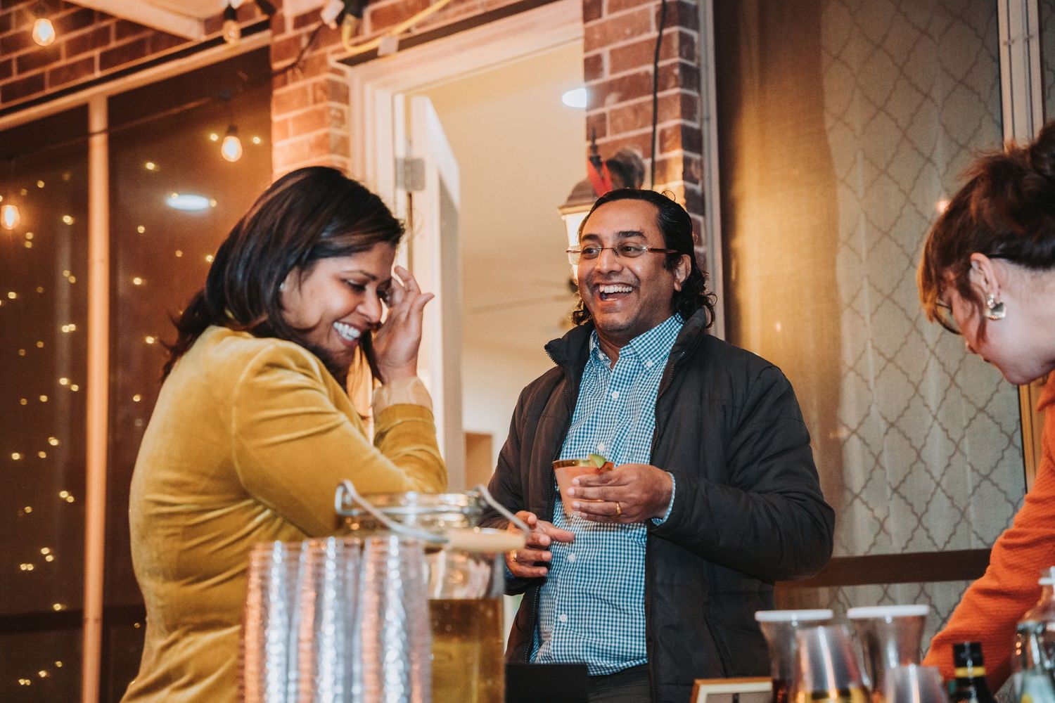 People sharing laughs and drinks at an evening party with string lights in background.