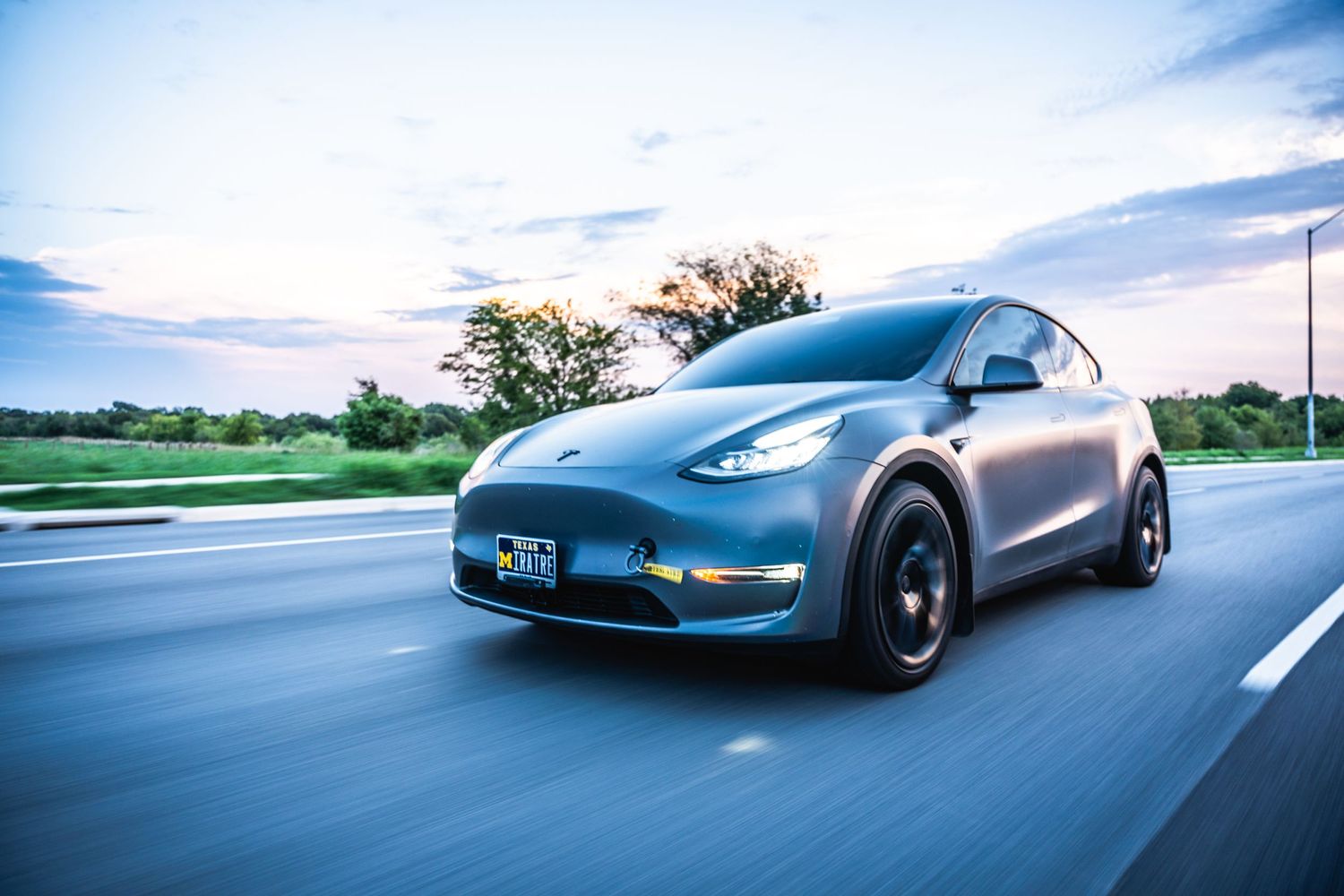 Matte gray Tesla Model Y driving on highway under blue skies with motion blur effect.