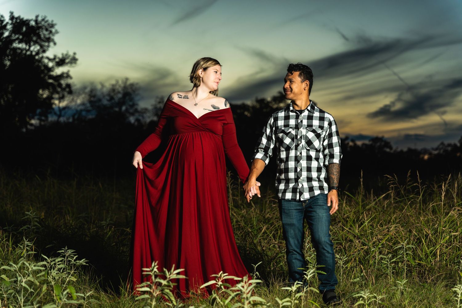 A couple stands together at sunset, with the woman wearing a long red dress in a grassy field.