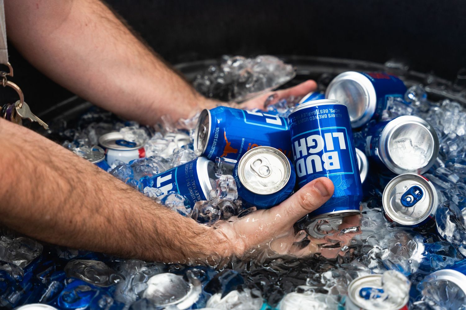 Hand reaching into cooler of ice to grab Bud Light cans at outdoor event.