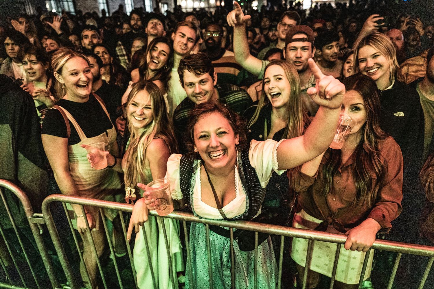 Enthusiastic concert crowd leans against barrier in green-lit venue during live music performance.