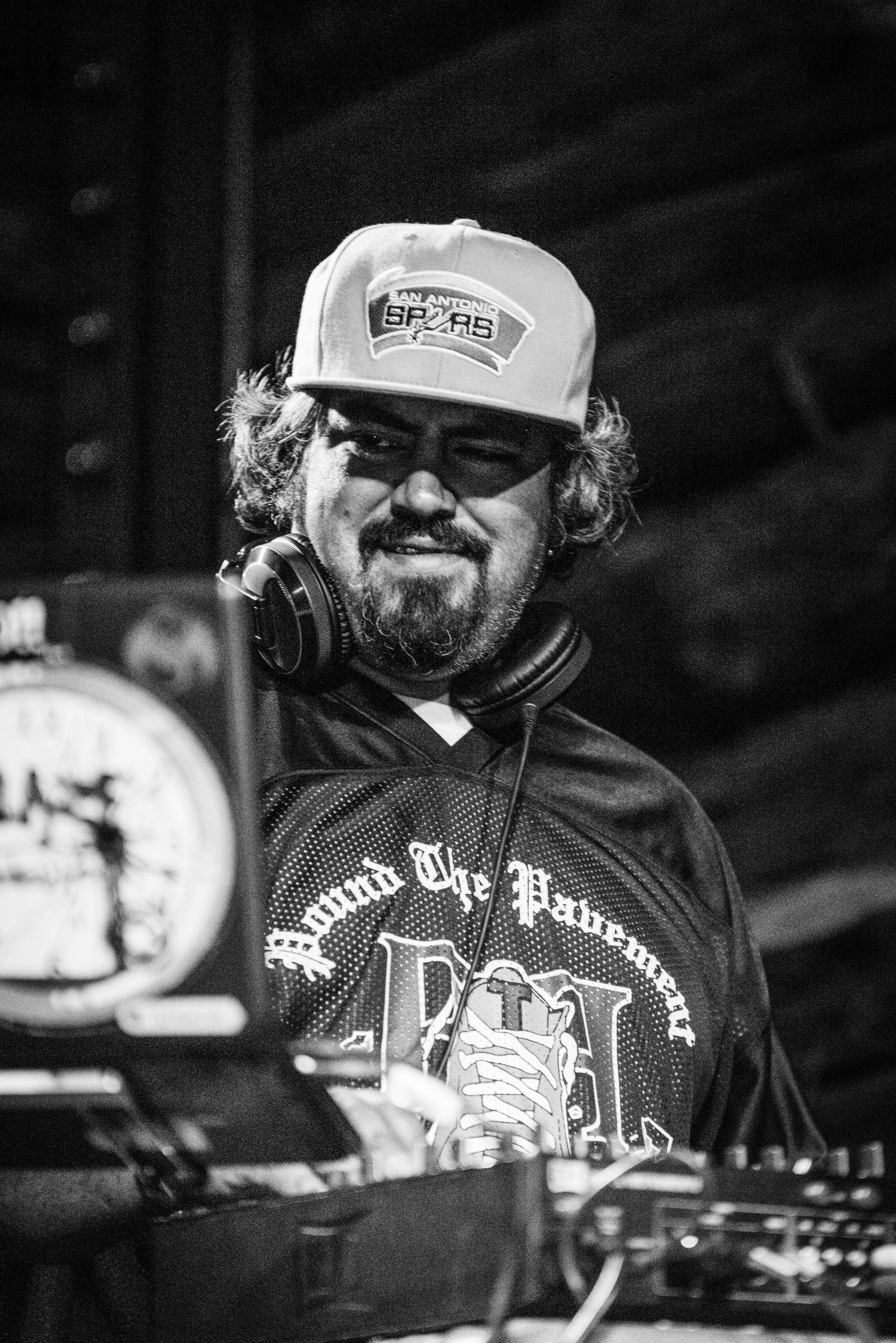 DJ wearing baseball cap and headphones smiles while performing at mixing deck in black and white photo.
