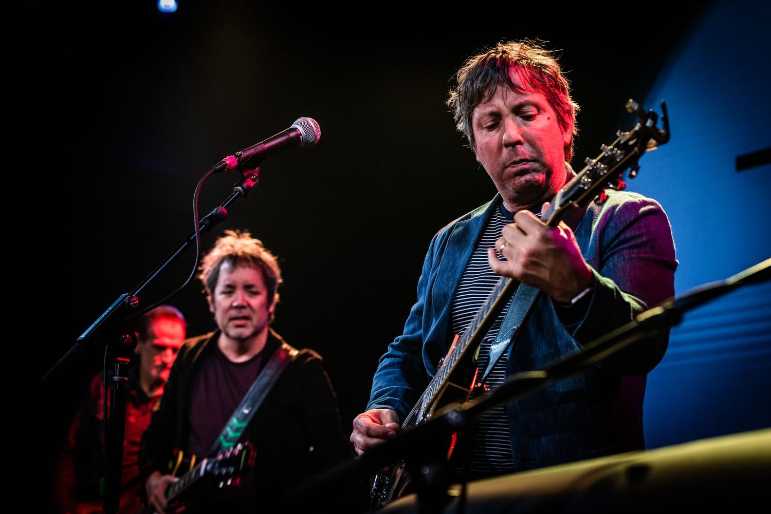 Musicians performing with electric guitars under colorful stage lighting during live show.