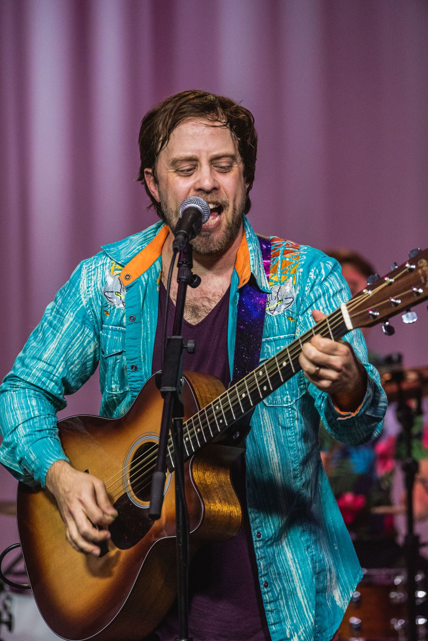 Musician in vibrant turquoise jacket plays sunburst acoustic guitar during performance.
