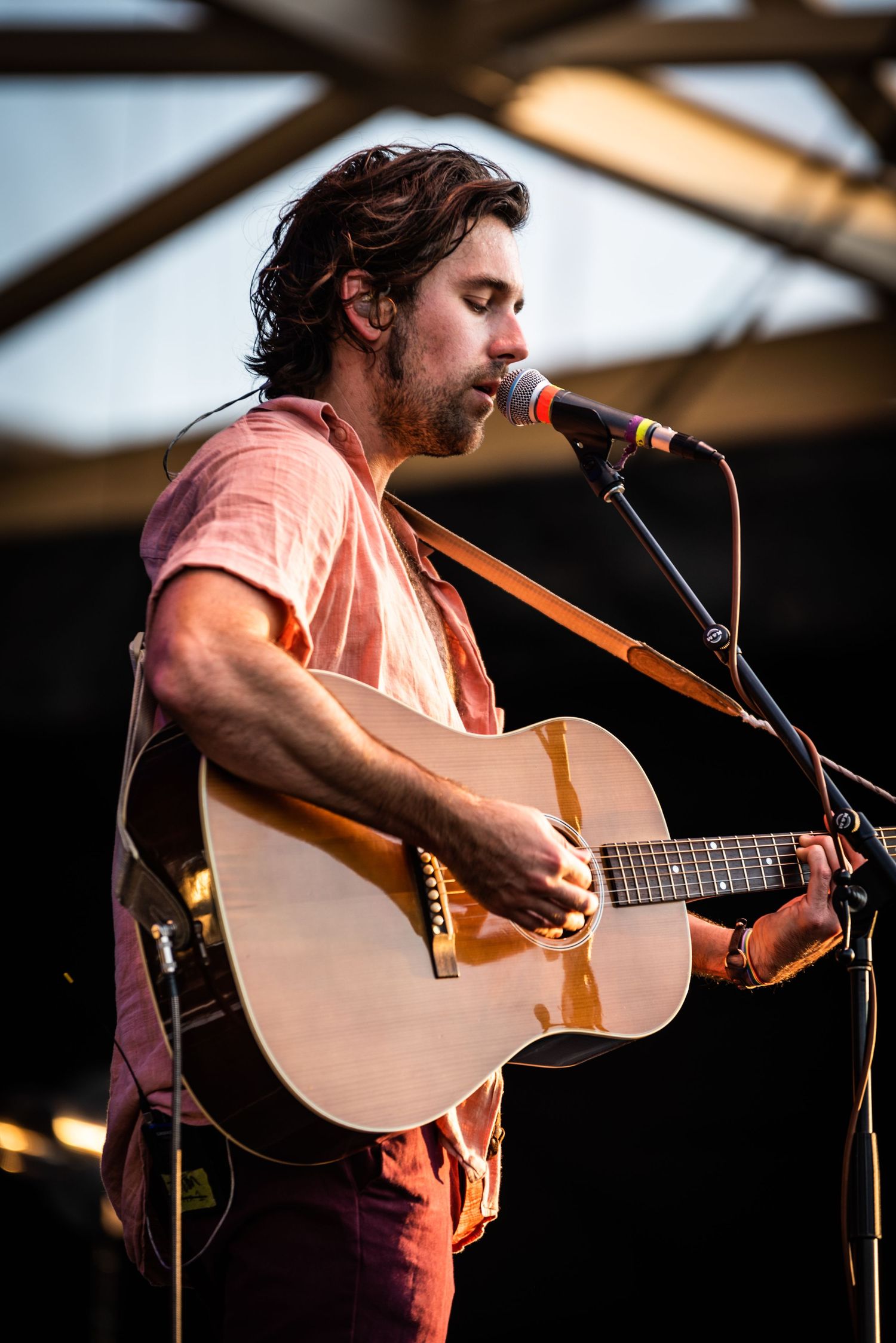 Musician performs with acoustic guitar wearing salmon colored shirt on outdoor stage.