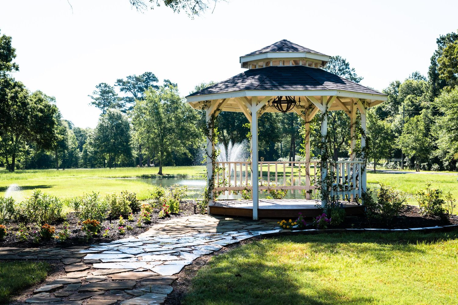 A white gazebo stands in a sunny park garden with a stone pathway leading to it.