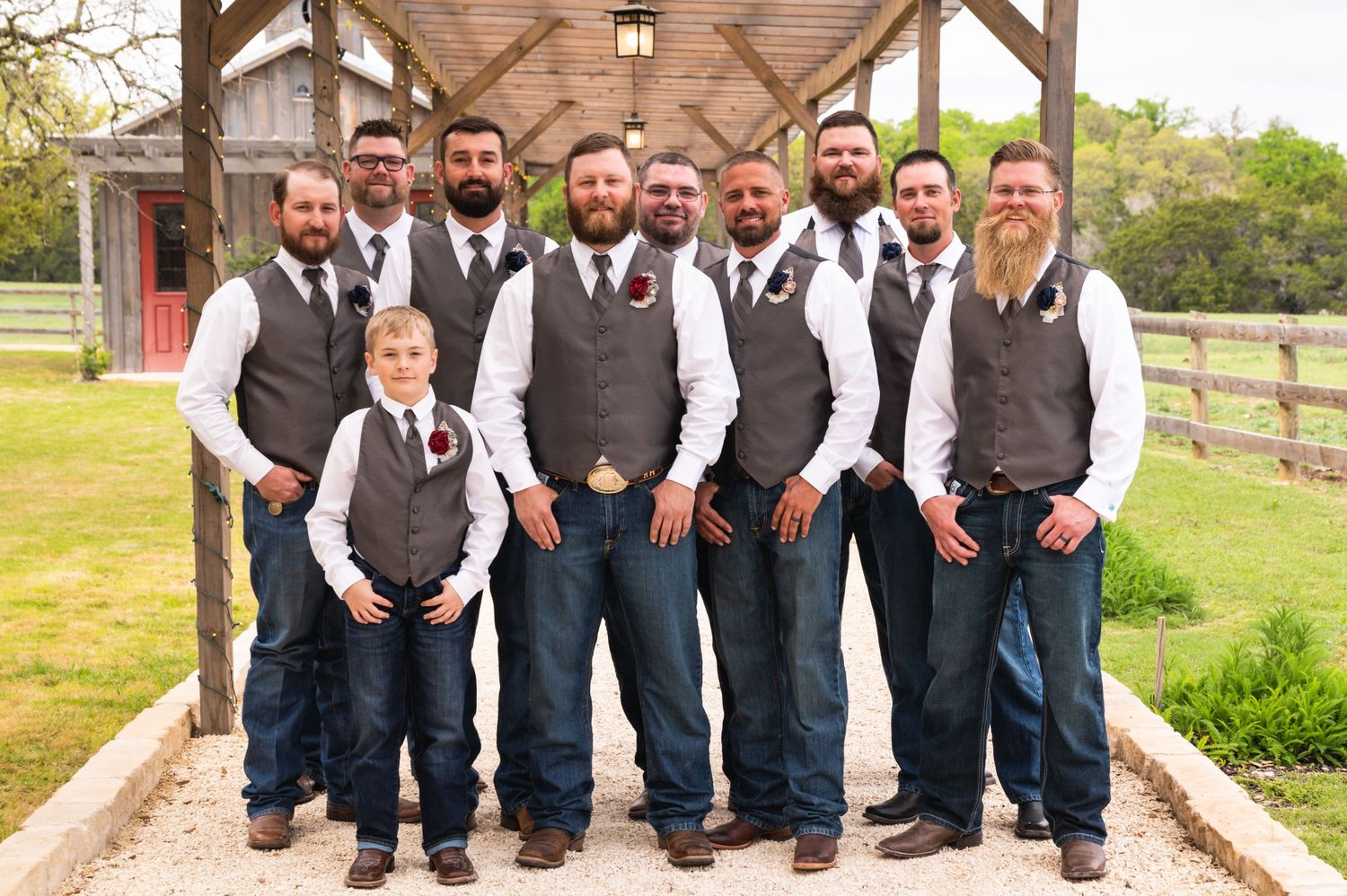 Groomsmen in coordinated grey vests and jeans pose together under a wooden pavilion.