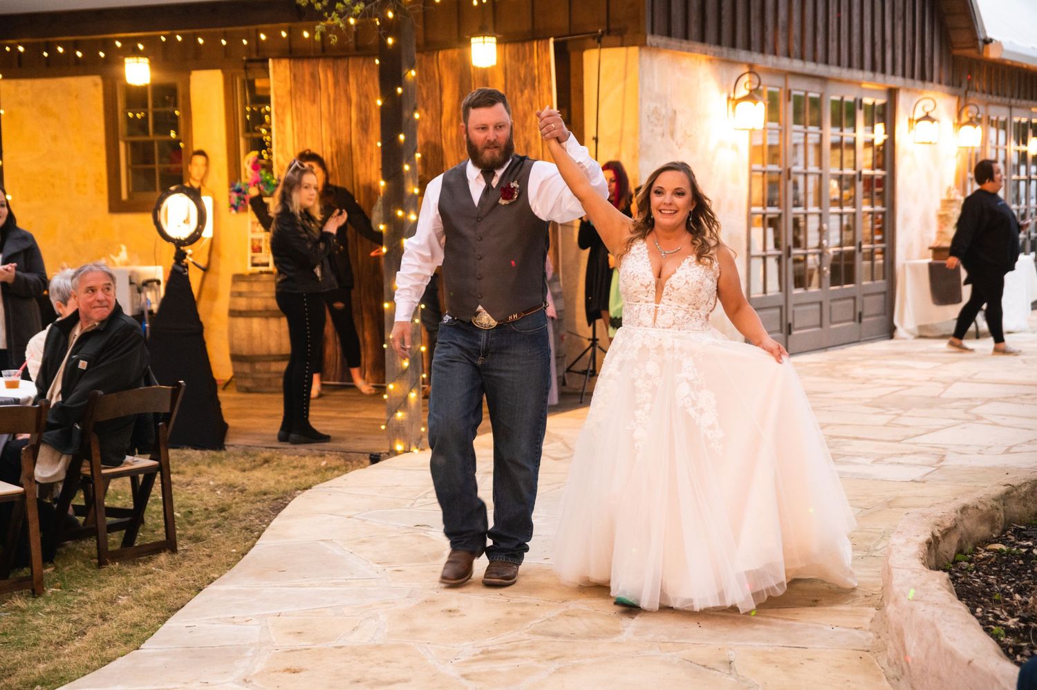 Newlyweds make their grand entrance at evening reception with string lights and venue buildings visible.