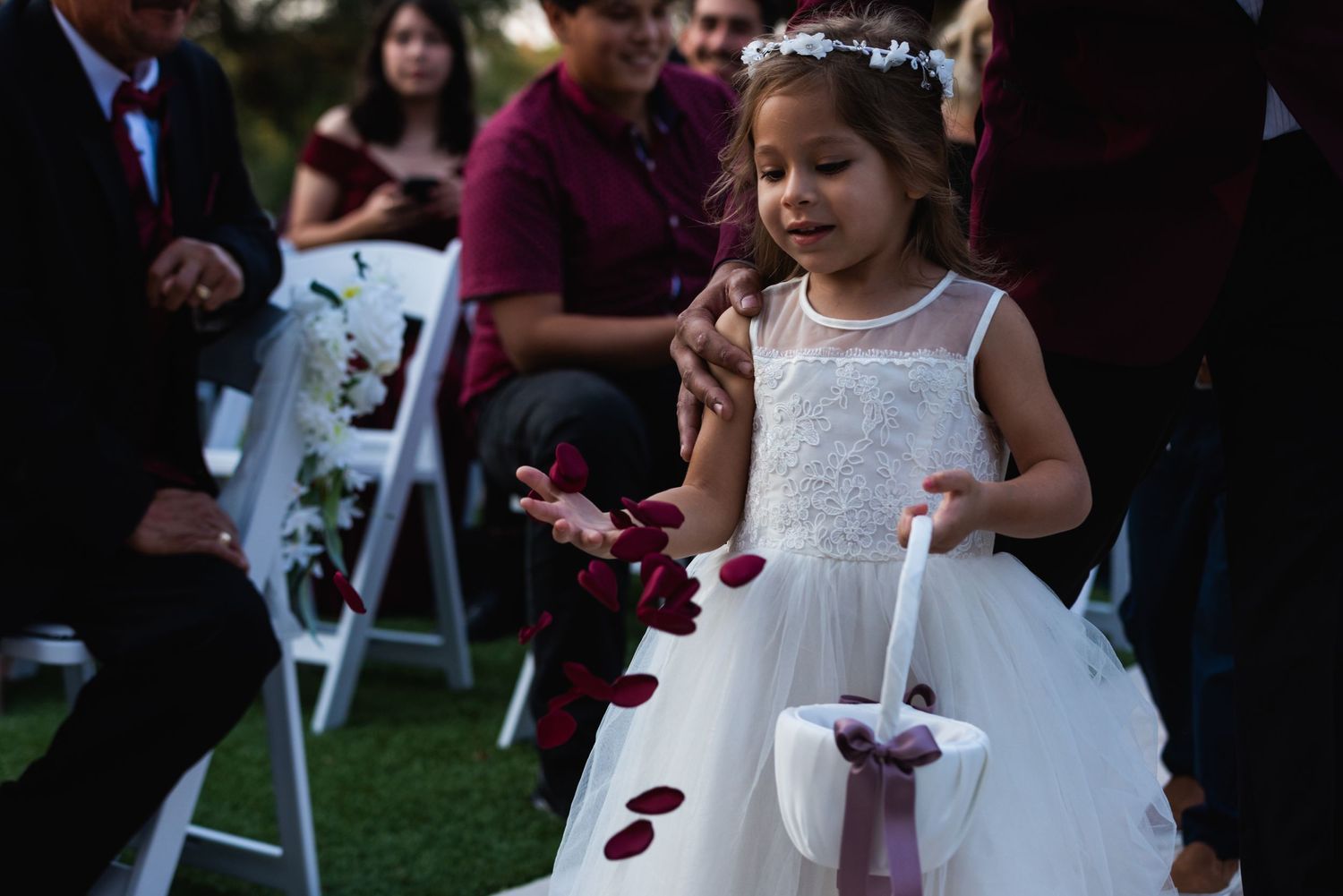 Flower girl in white dress scatters petals along wedding ceremony path.